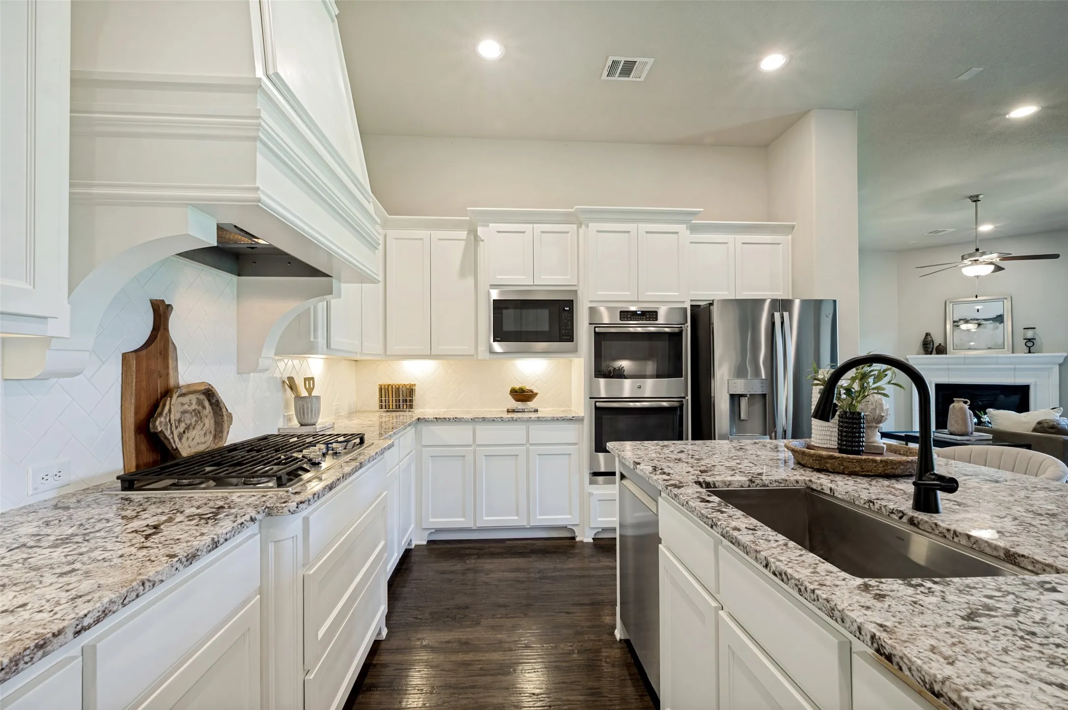 A beautifully appointed chef’s kitchen pairs timeless elegance with everyday function, featuring ceiling-height custom cabinetry, granite countertops, double ovens, a gas cooktop, and a statement vent hood framed by designer tile. The oversized island with seating and prep sink anchors the space, while the open-concept layout flows seamlessly into the living and dining areas for effortless entertaining.
