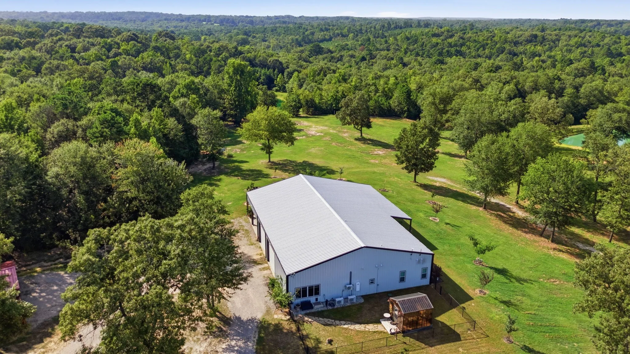 Expansive green space with mature trees, a metal-roof structure, and surrounding wooded areas