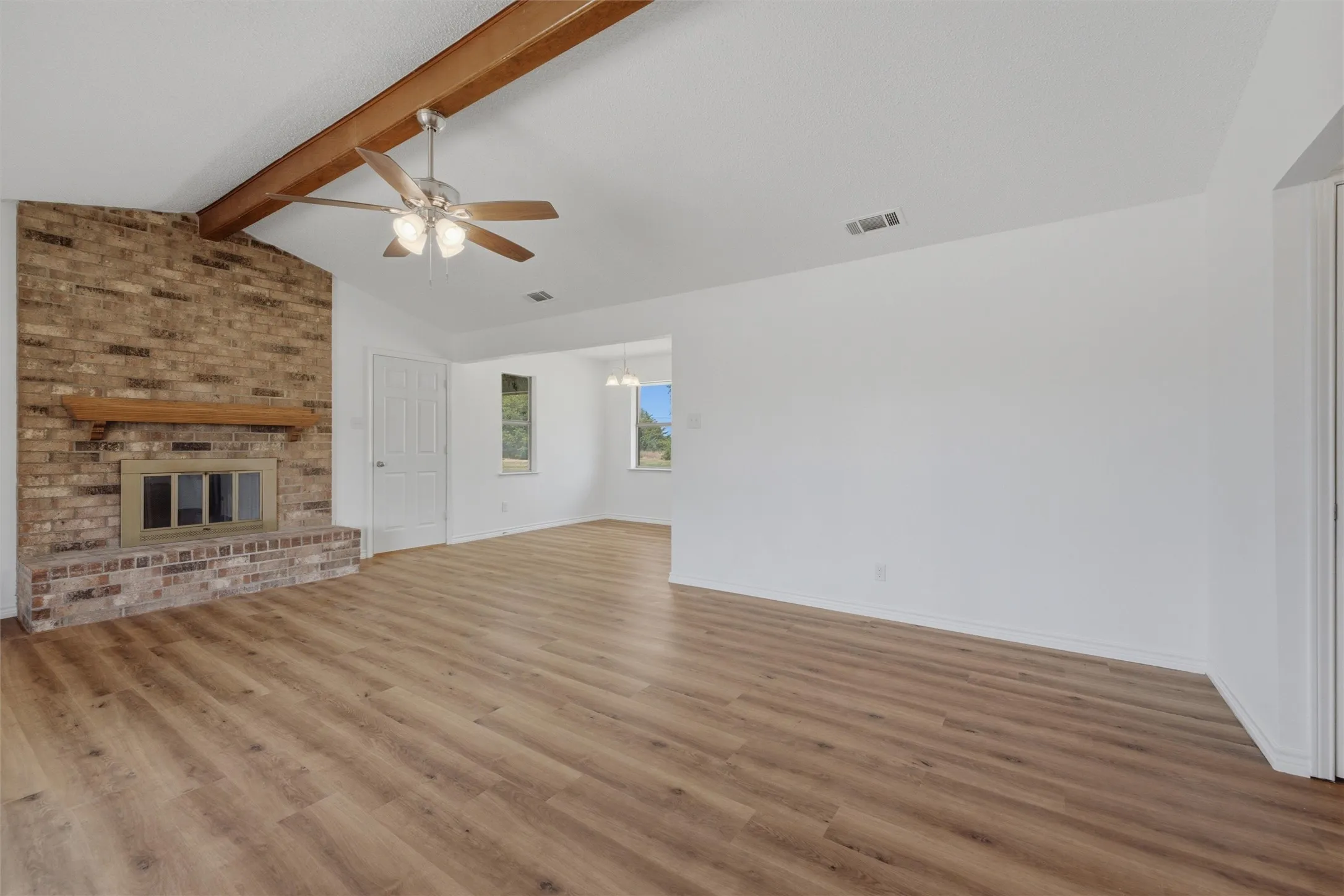 Vaulted ceiling with exposed wood beam, wood-finish flooring, brick fireplace with wood mantel, ceiling fan with light kit, and multiple windows