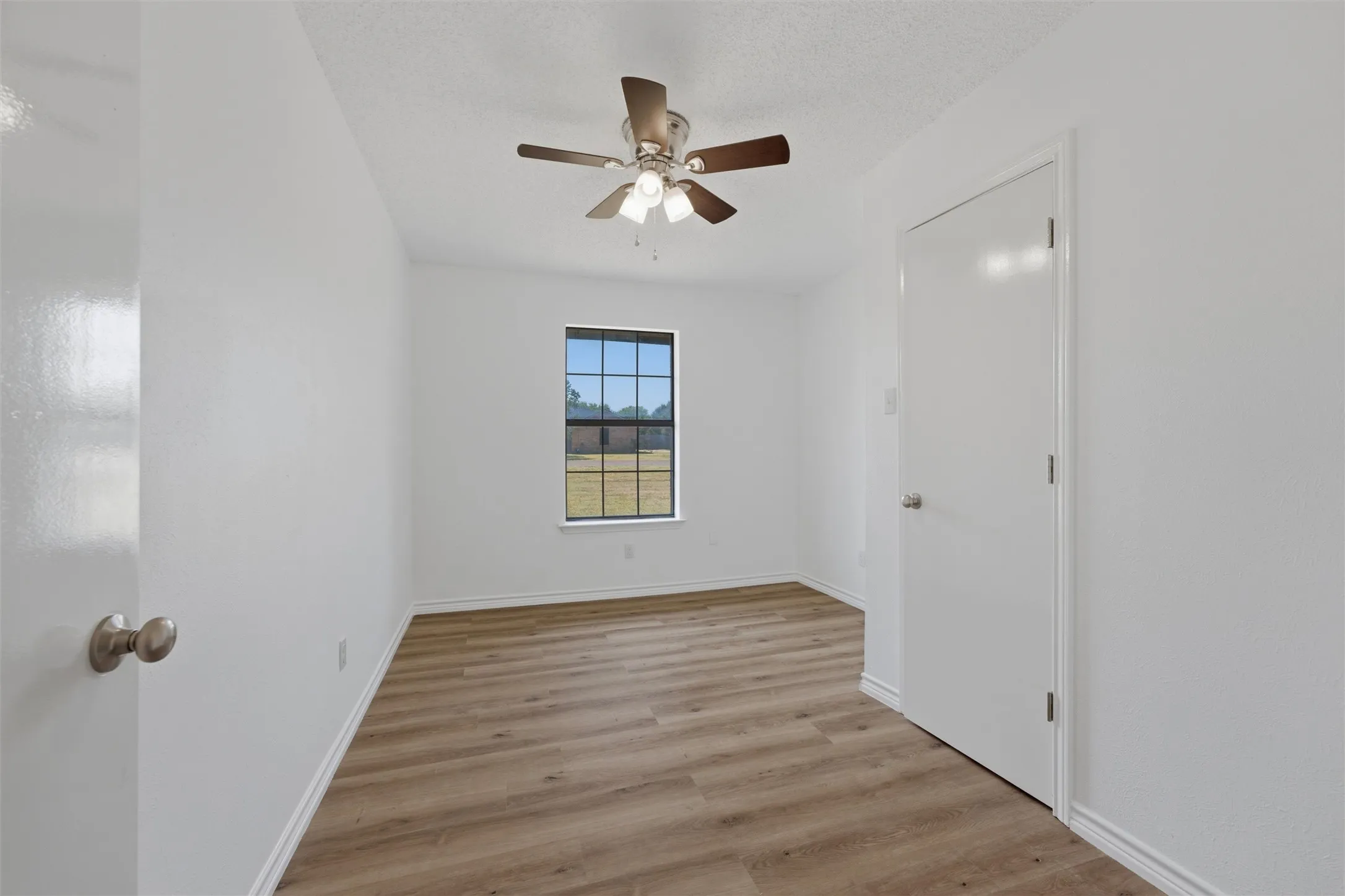 Wood-finish flooring, white walls, ceiling fan with integrated lighting, and a single window with dark framing