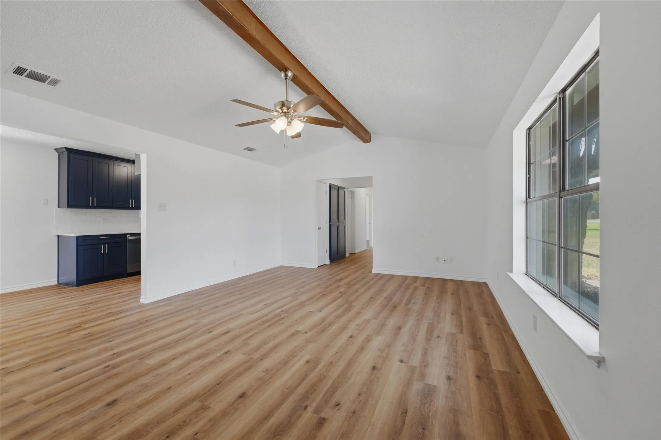 Vaulted ceiling with exposed wood beam, wood-finish flooring, ceiling fan with integrated lighting, and a large multi-pane window
