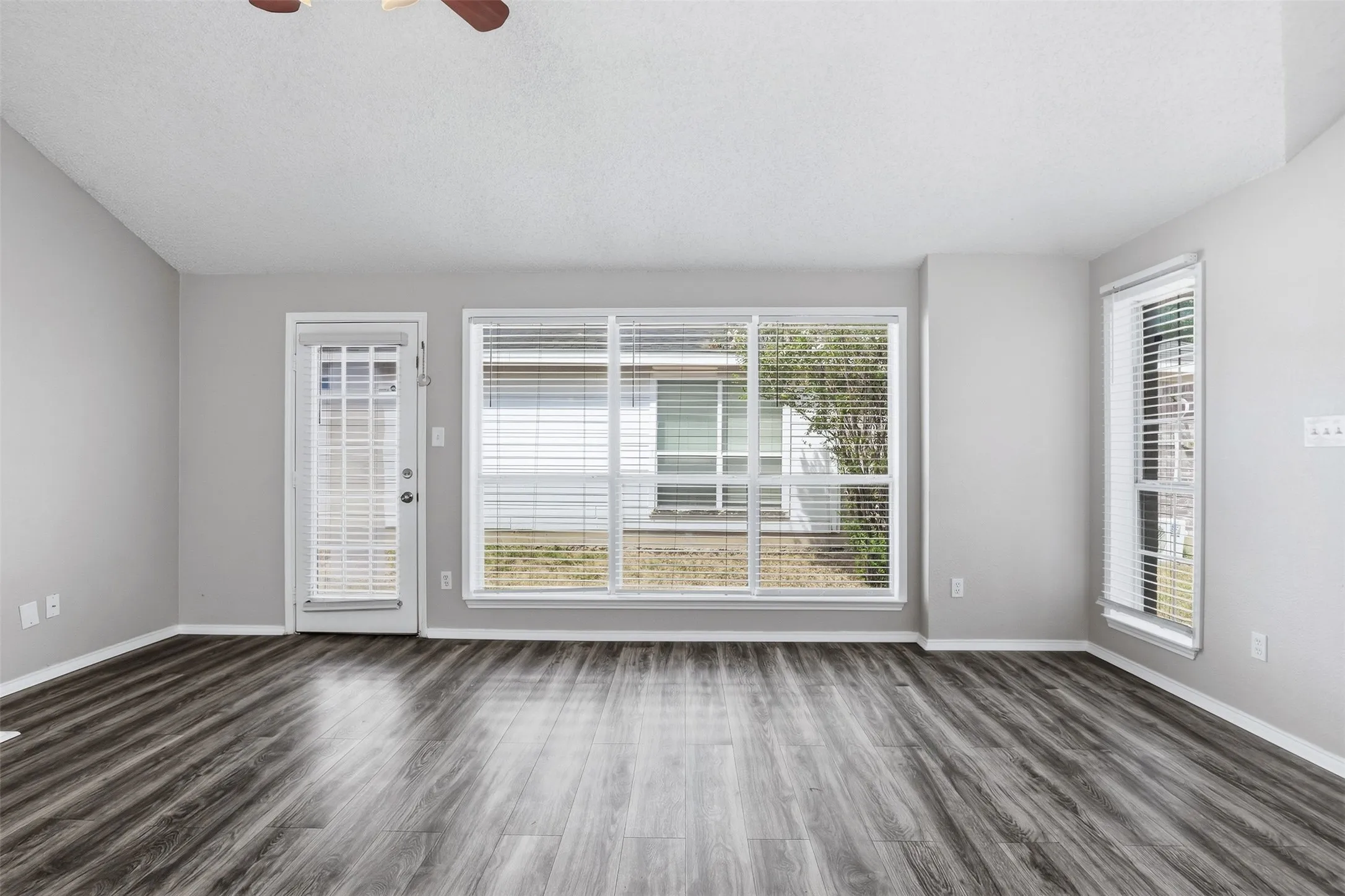 Spacious room featuring wood-finish flooring, multiple windows with blinds, a glass-paneled exterior door, and a ceiling fan