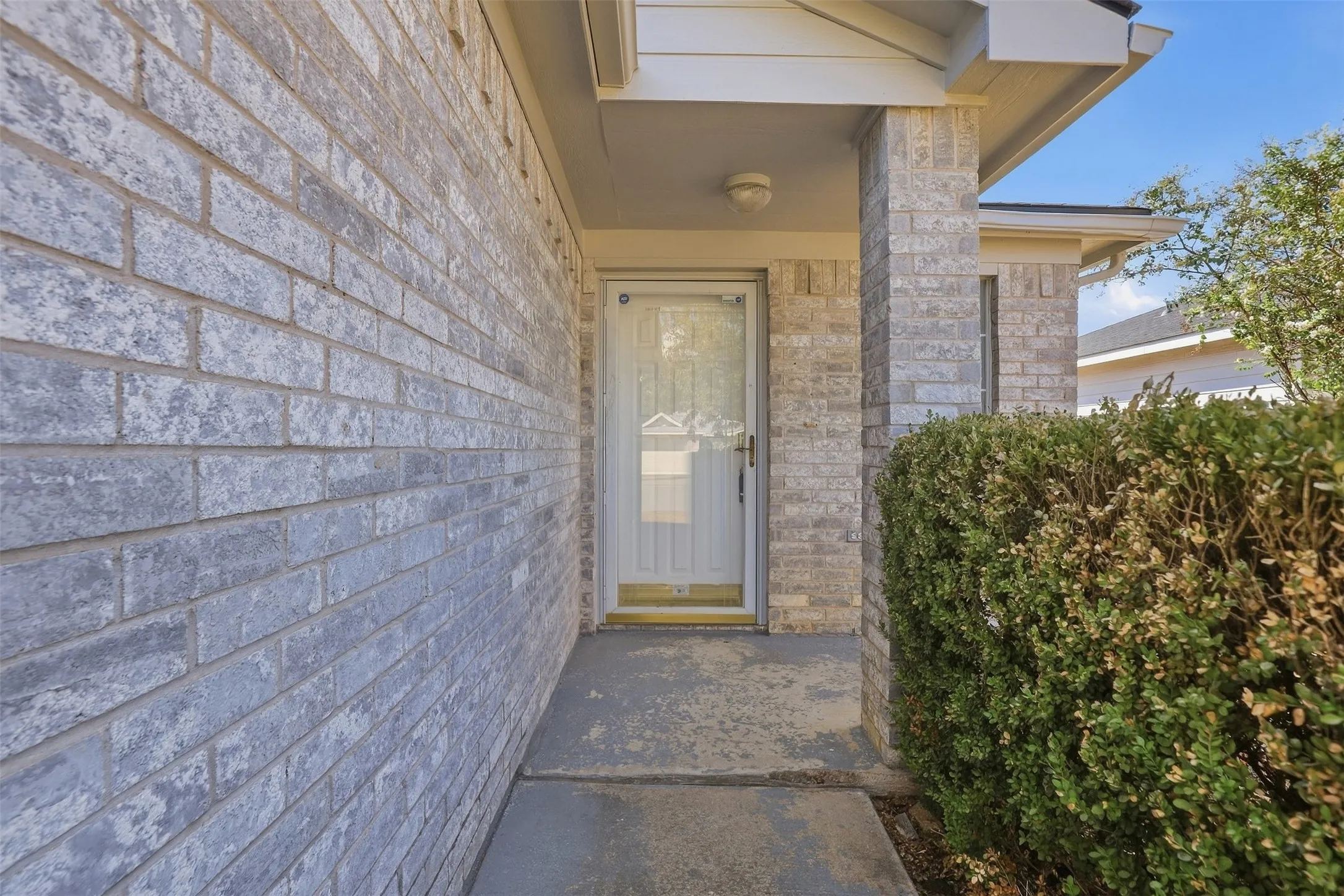 Brick exterior with a covered entryway, concrete pathway, and mature shrubbery