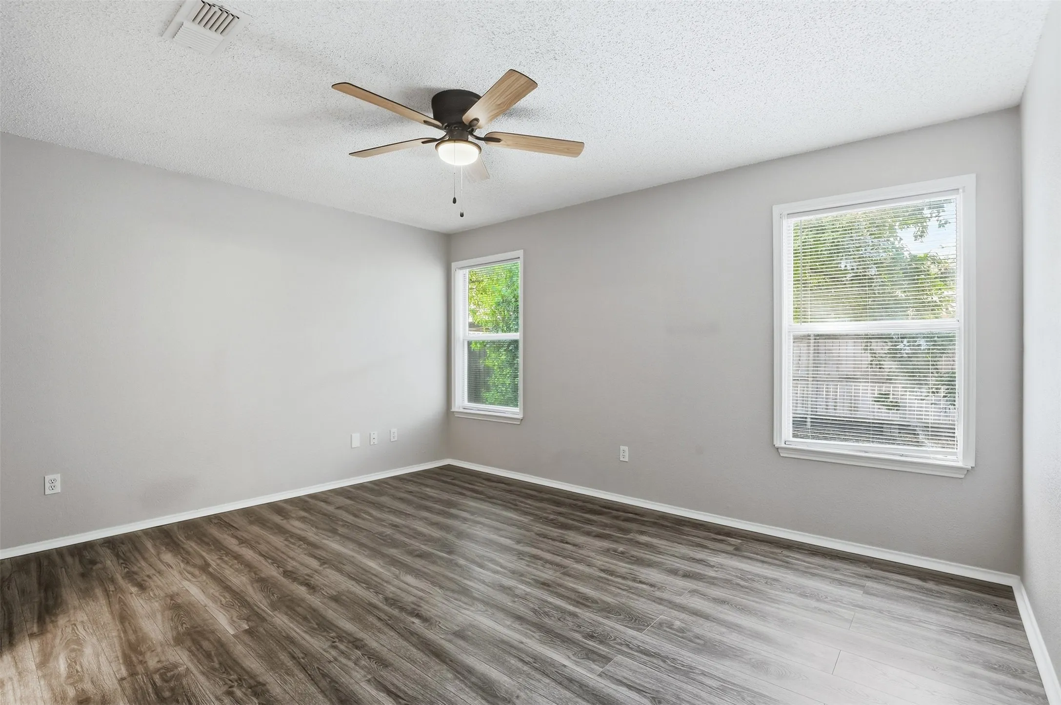 Wood-finish flooring, light gray walls, two windows with white trim, and a ceiling fan with integrated lighting