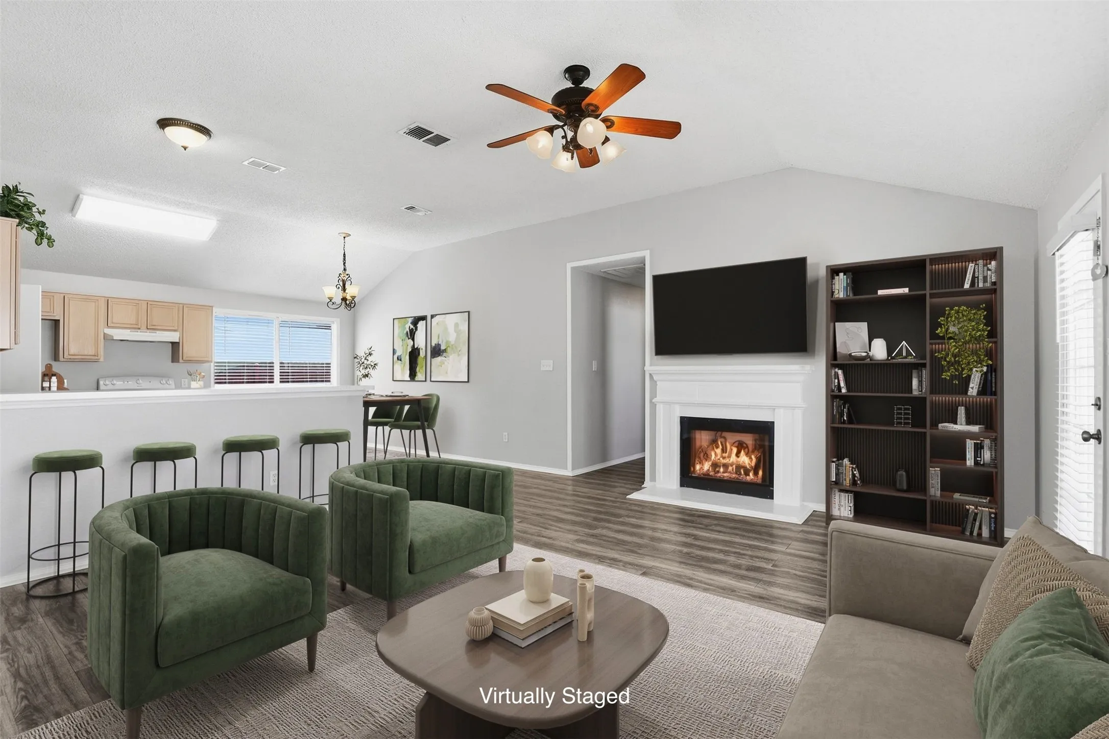 Vaulted ceiling with a ceiling fan, wood-finish flooring, built-in decorative fireplace with a white mantel, kitchen bar with a light wood-finish cabinetry, and a built-in bookshelf