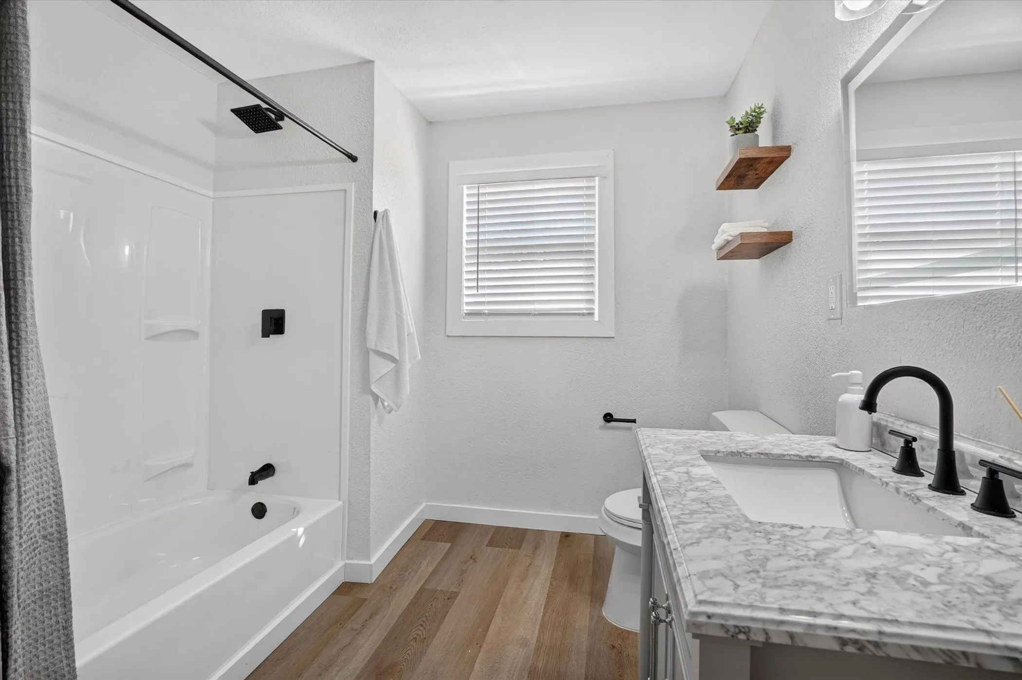Bathroom featuring wood-finish flooring, a white tub with a black rain shower head, a window with blinds, and a vanity with a white countertop and black fixtures