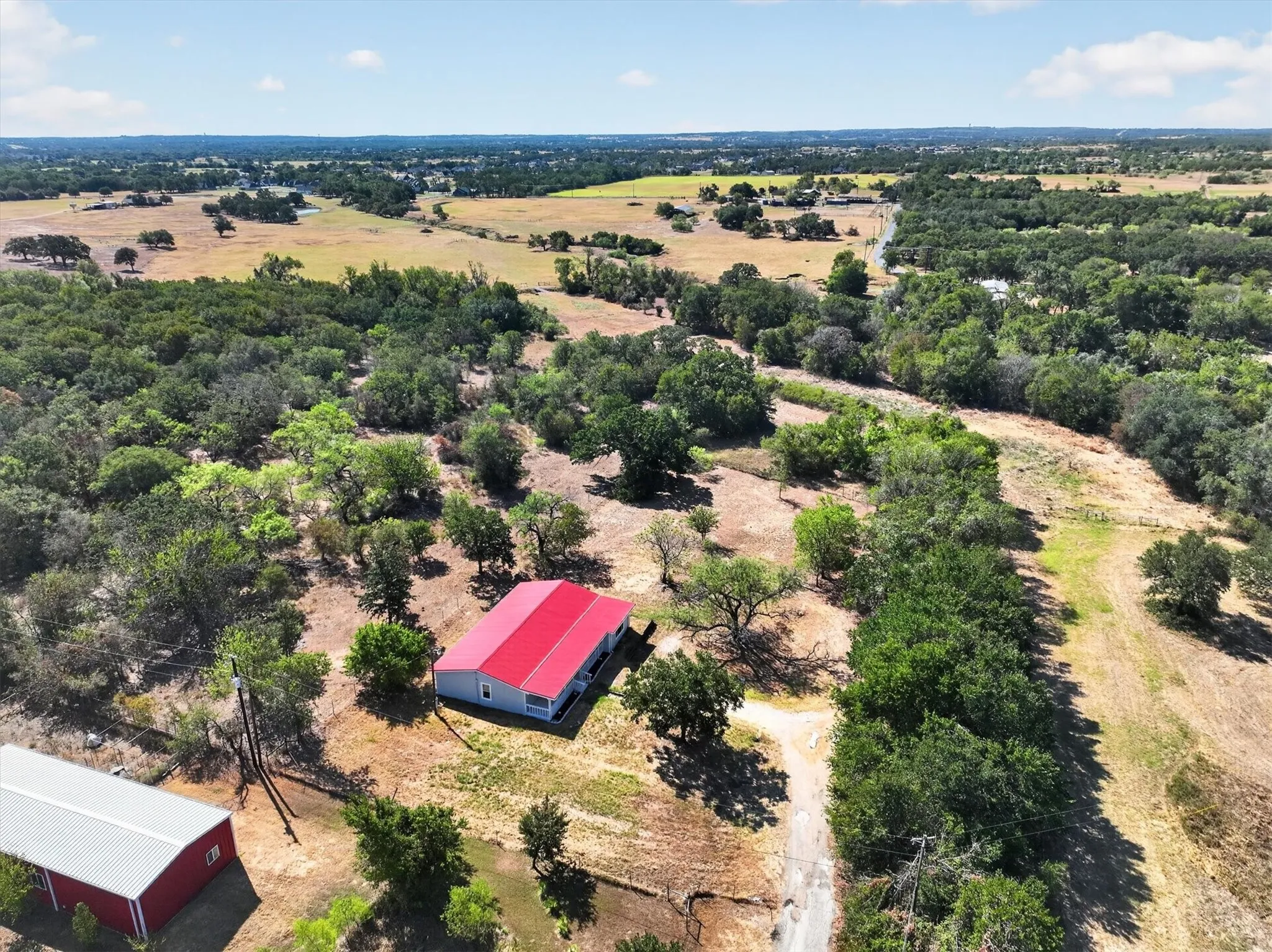 Expansive land featuring a main structure with a red metal roof, a secondary red barn-style structure with a metal roof, and scattered mature trees