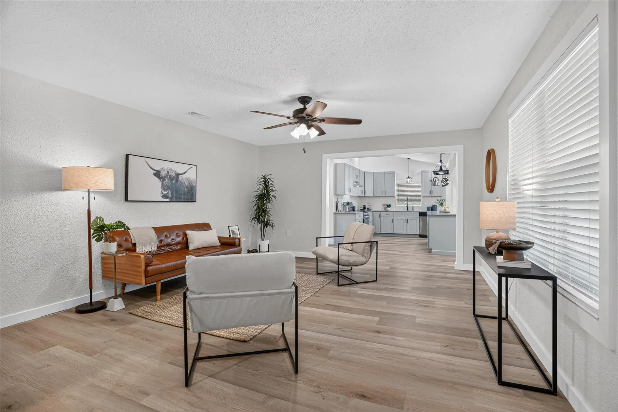 Wood-finish flooring throughout, ceiling fan with light fixture, ample natural light from multiple windows, and a clear architectural opening to the kitchen