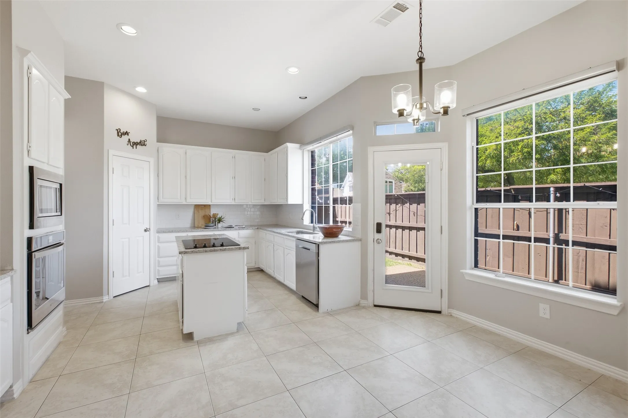 A thoughtfully laid-out kitchen with white raised-panel cabinetry, granite countertops, a center island with cooktop, stainless steel appliances, and an eat-in breakfast area bathed in natural light from a large picture window and a glass back door.
