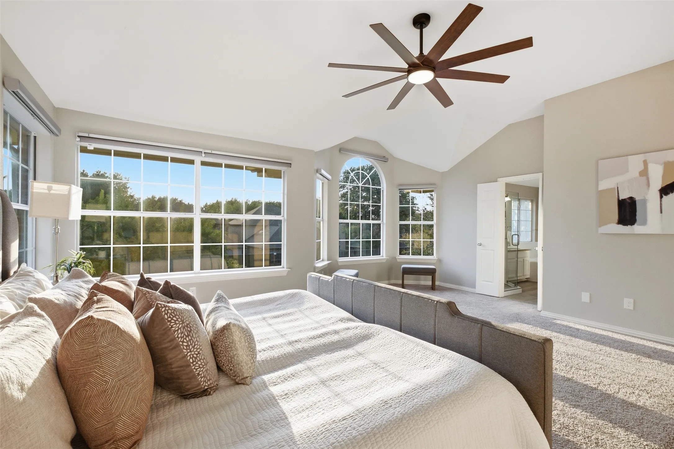 Sunlight pours through the arched and gridded windows of the primary bedroom, highlighting the vaulted ceiling and the generous proportions of this rear-facing retreat. The entry to the primary bathroom is visible in the background, completing the private suite layout.
