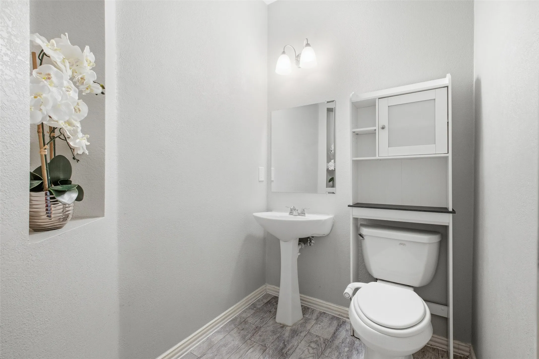 A clean, classic half bath on the main level features a pedestal sink, sconce lighting, framed mirror, and a decorative wall niche — everything guests need without any wasted space.