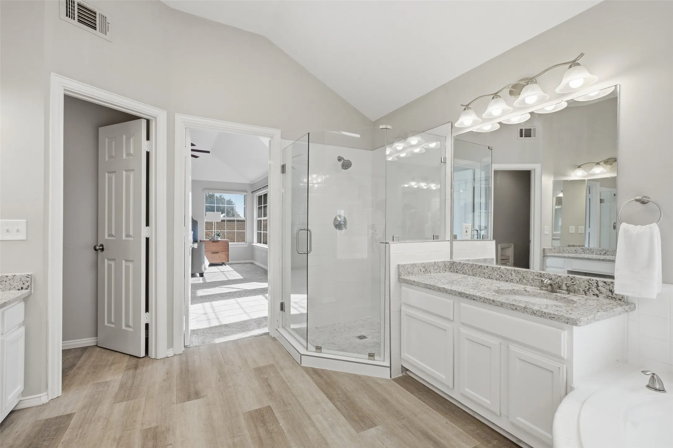 The glass-enclosed walk-in shower with white subway tile and pebble-tile floor is a standout feature of the primary bathroom. Granite vanities and LVP floors extend throughout, while the open door frames a view back into the primary bedroom.
