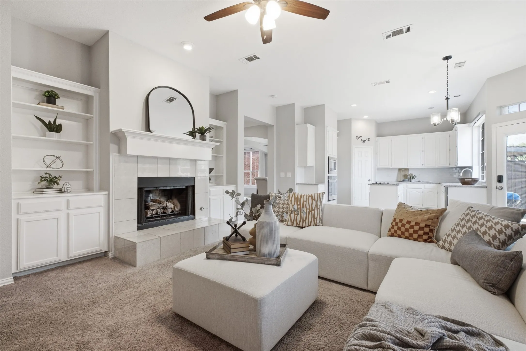 The heart of the home with soaring ceilings, a cozy tiled wood-burning fireplace framed by white built-in bookshelves and cabinetry, and a direct sightline into the kitchen. Neutral paint and plush carpet keep the space warm and inviting for everyday living or entertaining.