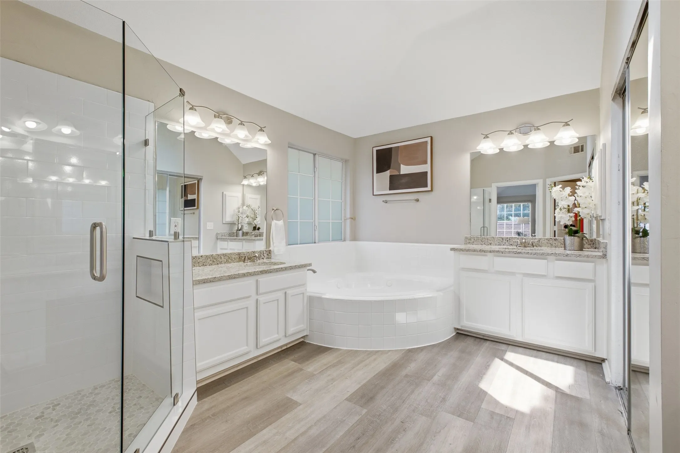 The primary bathroom delivers a true spa experience with dual granite-topped vanities, a large corner soaking tub with tile surround, a glass-enclosed walk-in shower, and updated LVP wood-look flooring. Two sets of sconce lighting keep the room bright morning and night.
