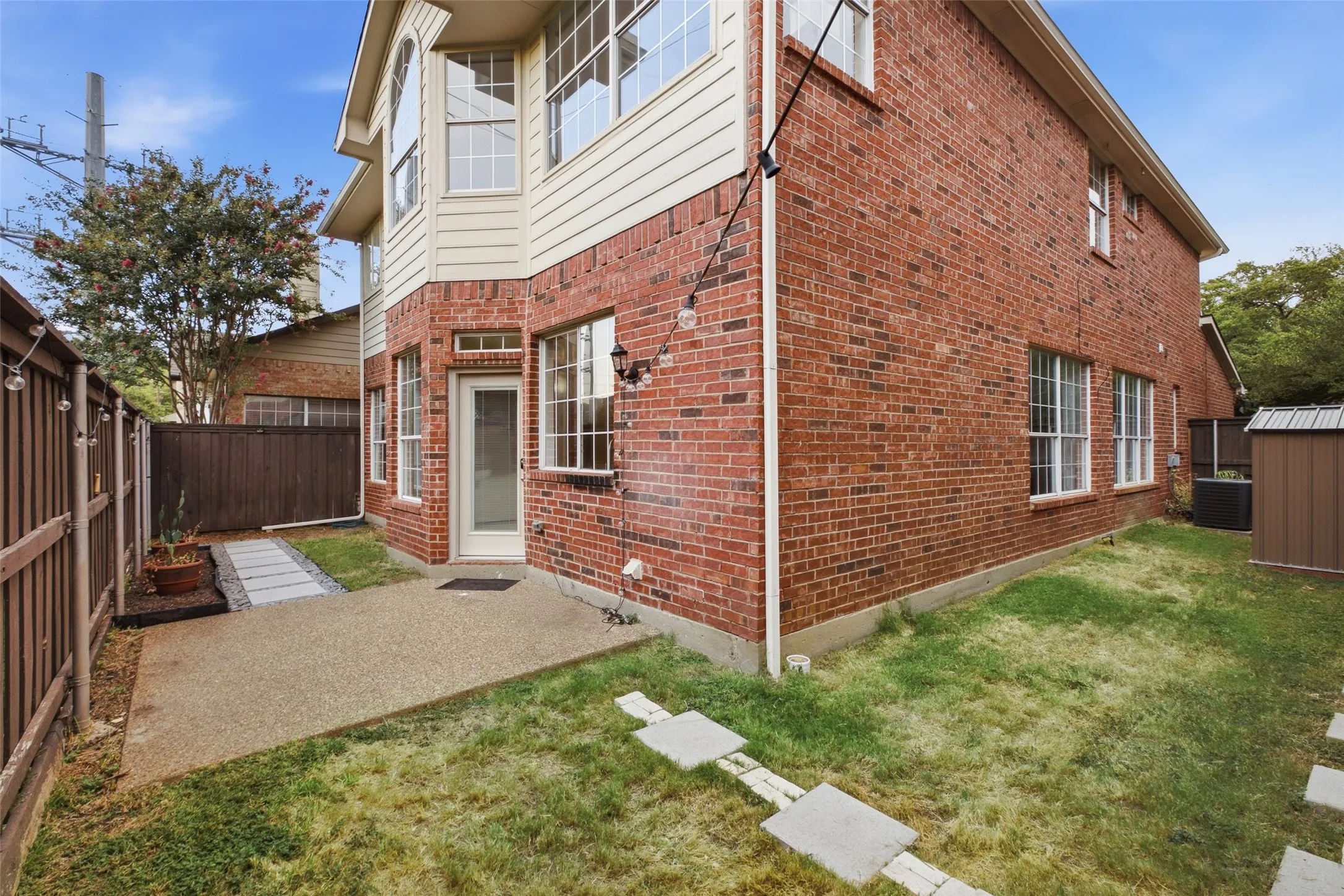 The private, fully fenced backyard features a concrete patio, green lawn, a charming side garden path with stepping stones, and a storage shed tucked into the corner. String lights and a blooming crape myrtle add welcoming character to the outdoor space.
