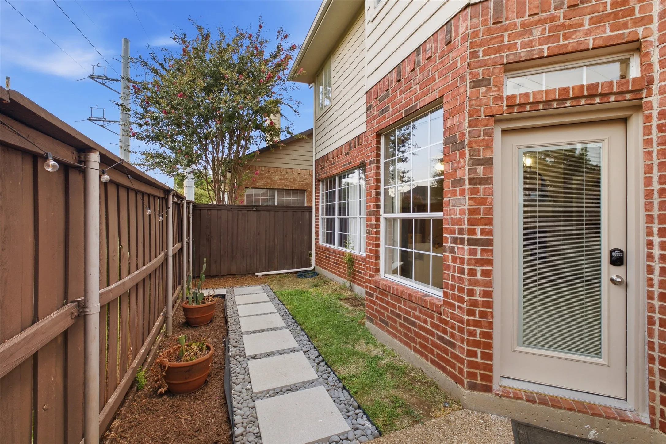 A beautifully detailed side yard with a stepping stone walkway edged in river rock, string lights strung along the fence, and a glass-panel back door with keypad entry. It's the kind of thoughtful outdoor detail that shows real care for the home.