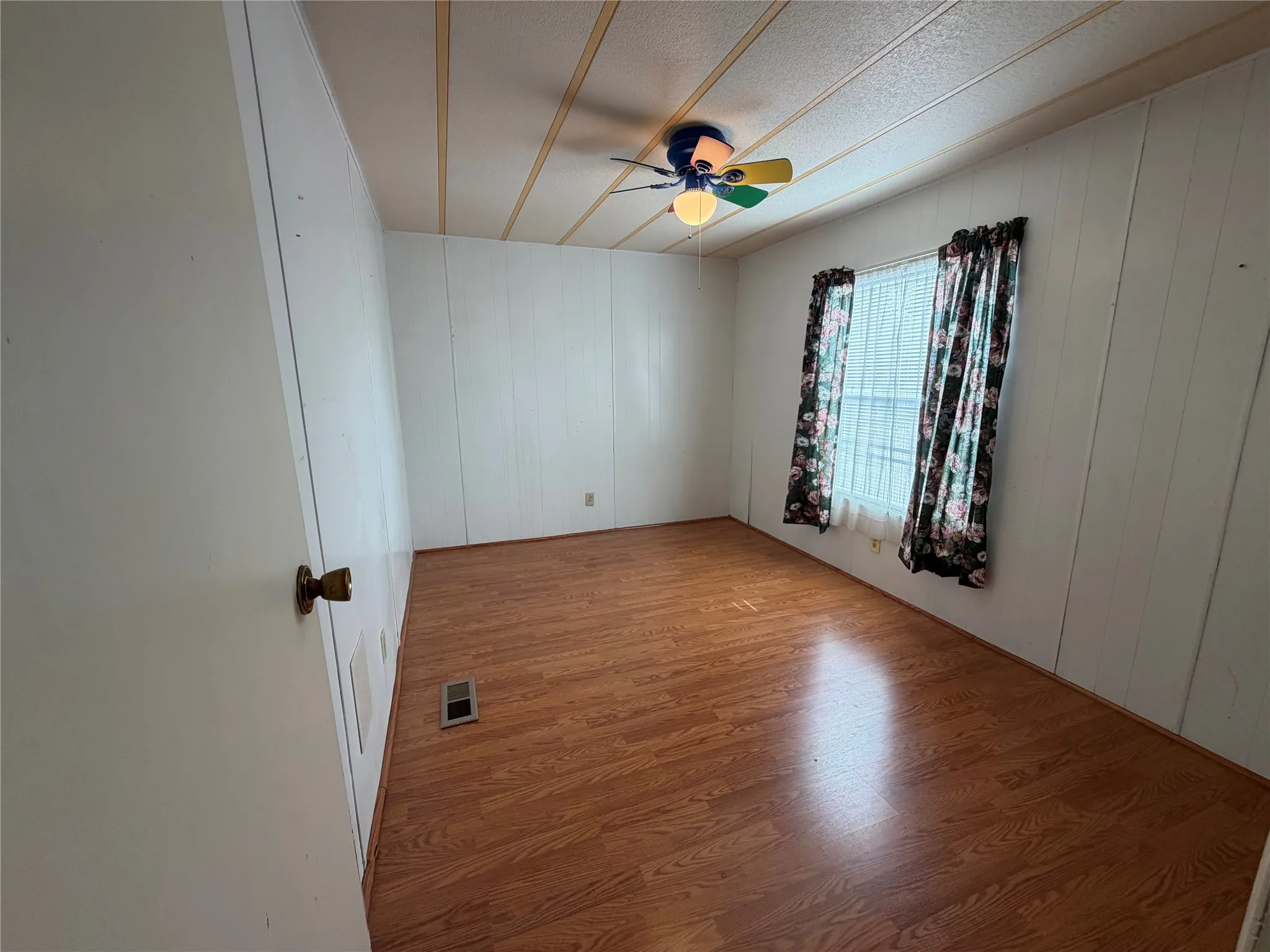 Spacious bedroom with warm wood-finish flooring, white wall paneling, ceiling-mounted fan with lighting fixture, and large window with patterned curtains allowing natural light