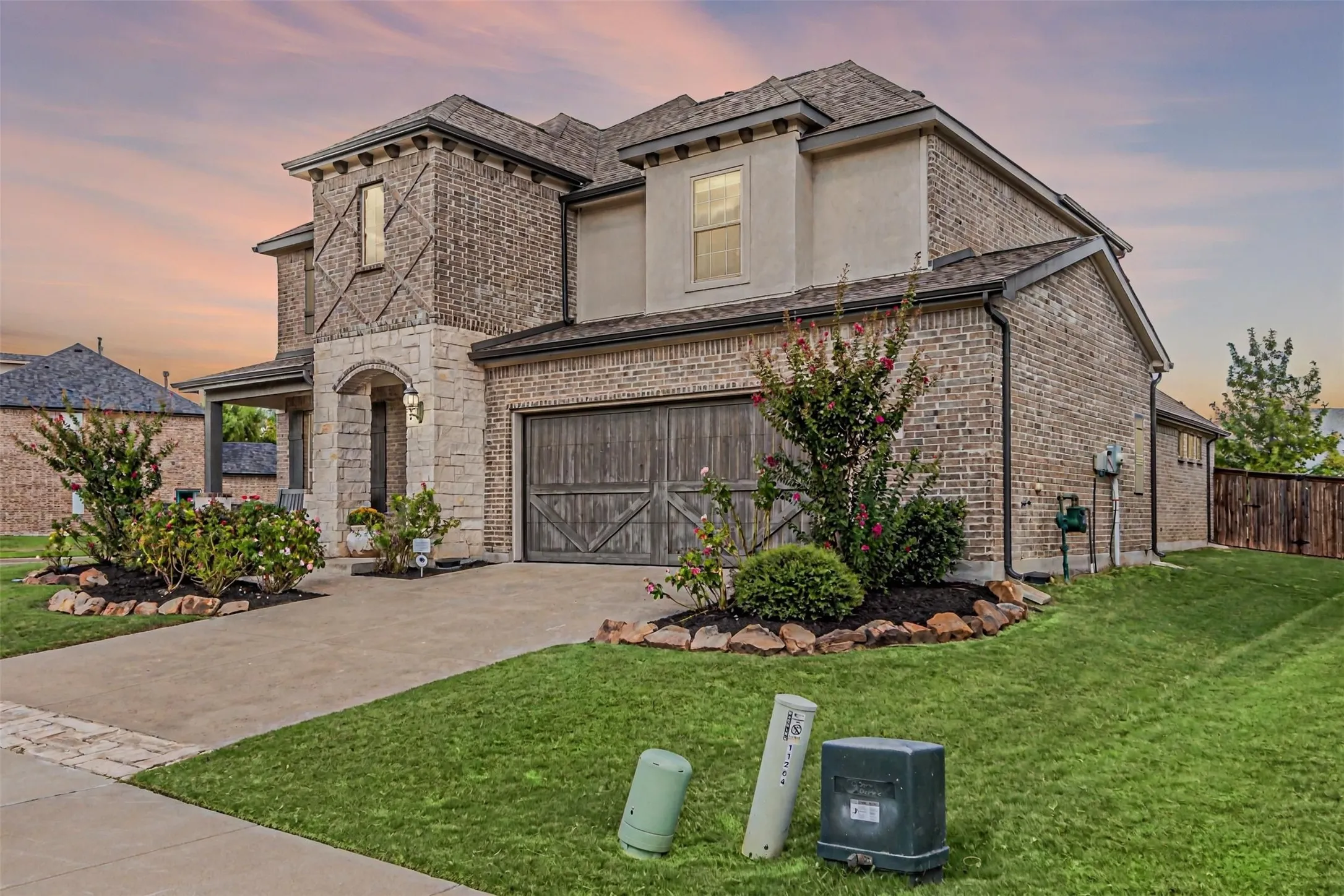 Brick and stone facade, arched entryway, wood-finish garage door, mature landscaping, and concrete driveway