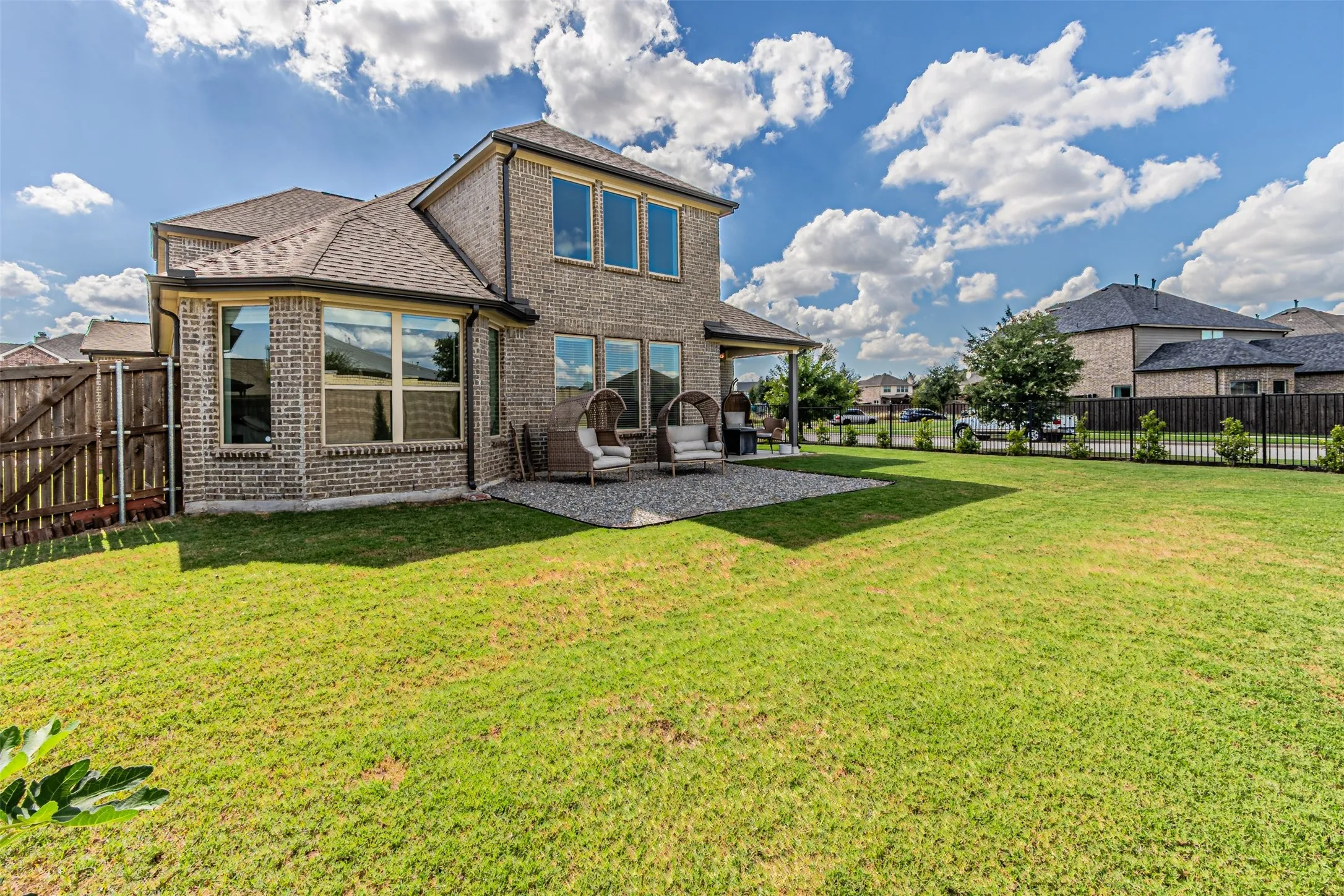 Expansive green lawn with a covered patio, a bay window, and a brick exterior, enclosed by a combination of wood and metal fencing