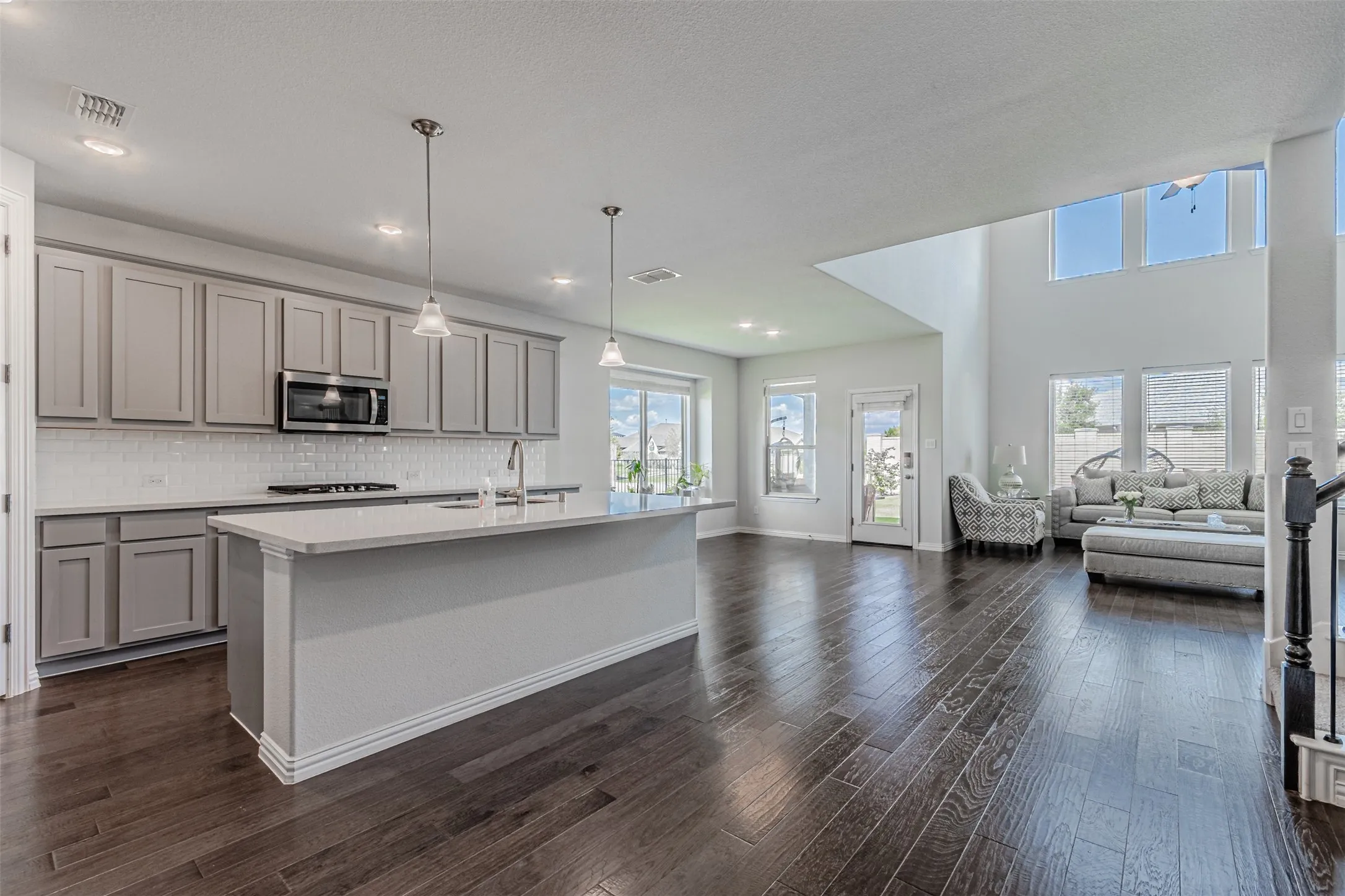 Open-concept living area featuring wood-finish flooring, a kitchen island with pendant lighting, stainless steel appliances, and a two-story ceiling with high windows