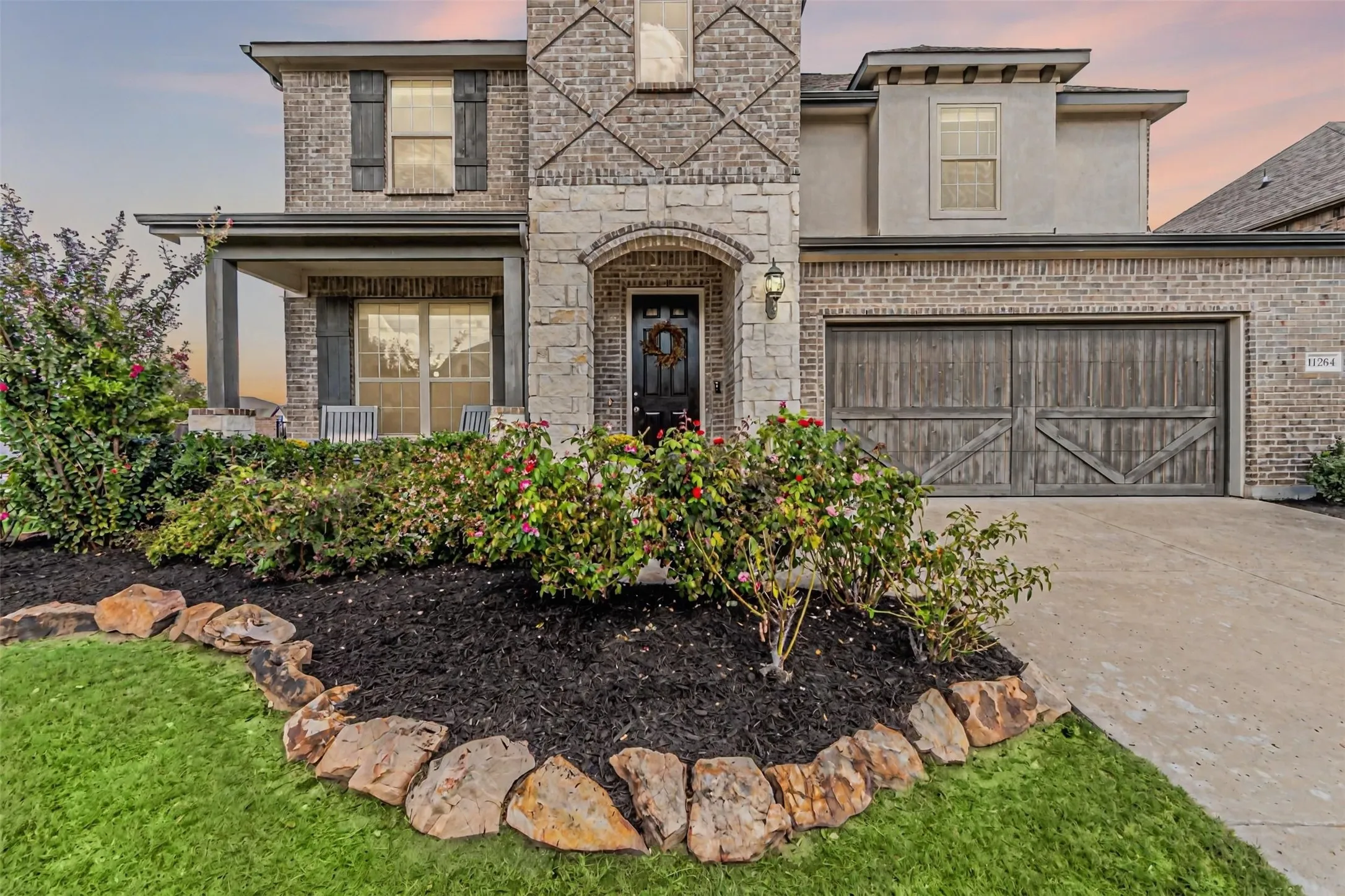 Brick and stone facade with a covered entry porch, dark wood-finish garage door, and a manicured front lawn with rock​​‌​​​​‌​​‌‌​‌‌​​​‌‌​‌​‌​‌​​​‌​​ edging