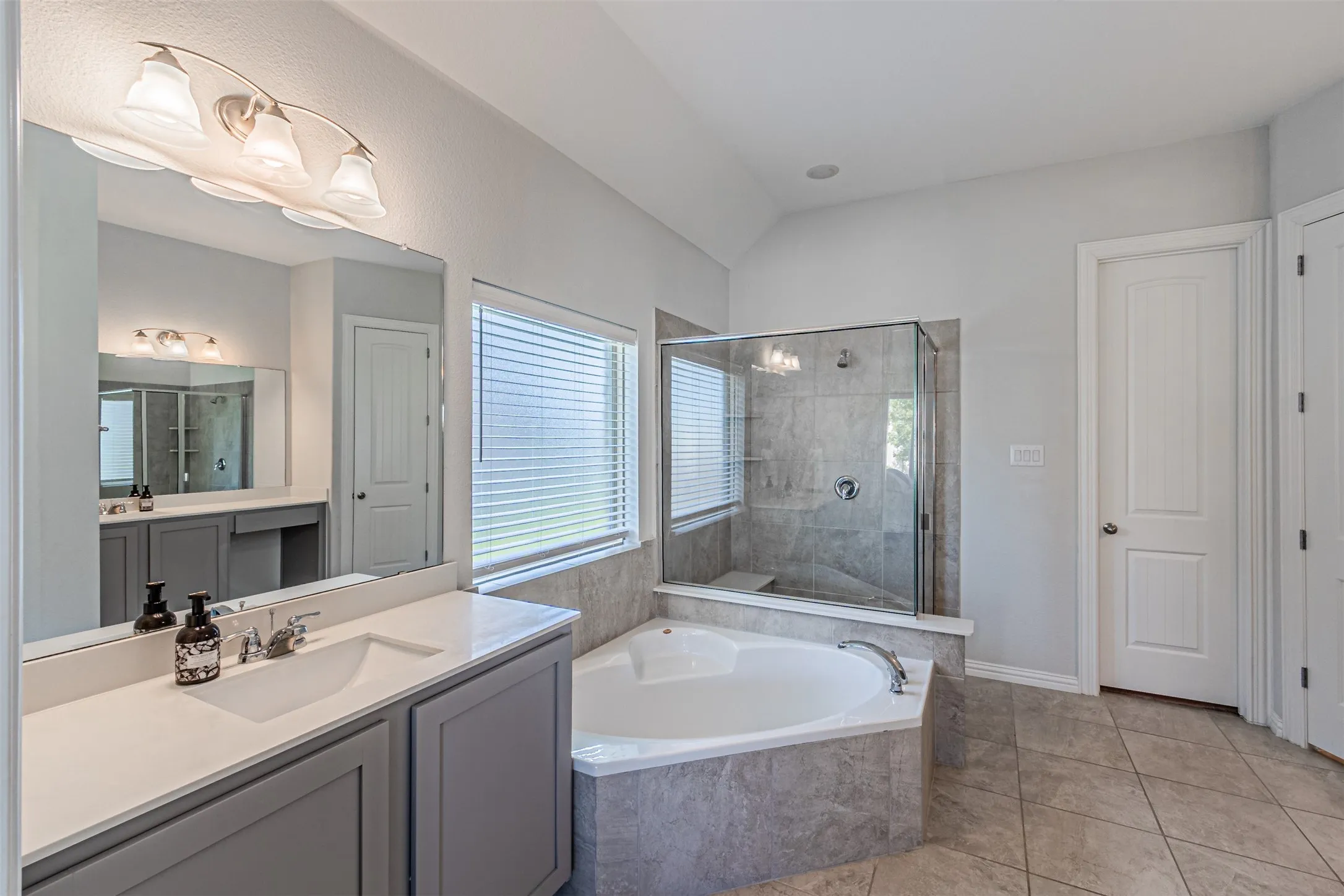 Dual vanity with gray cabinetry and white countertops, corner soaking tub, glass-enclosed shower with tile surround, and neutral tile flooring