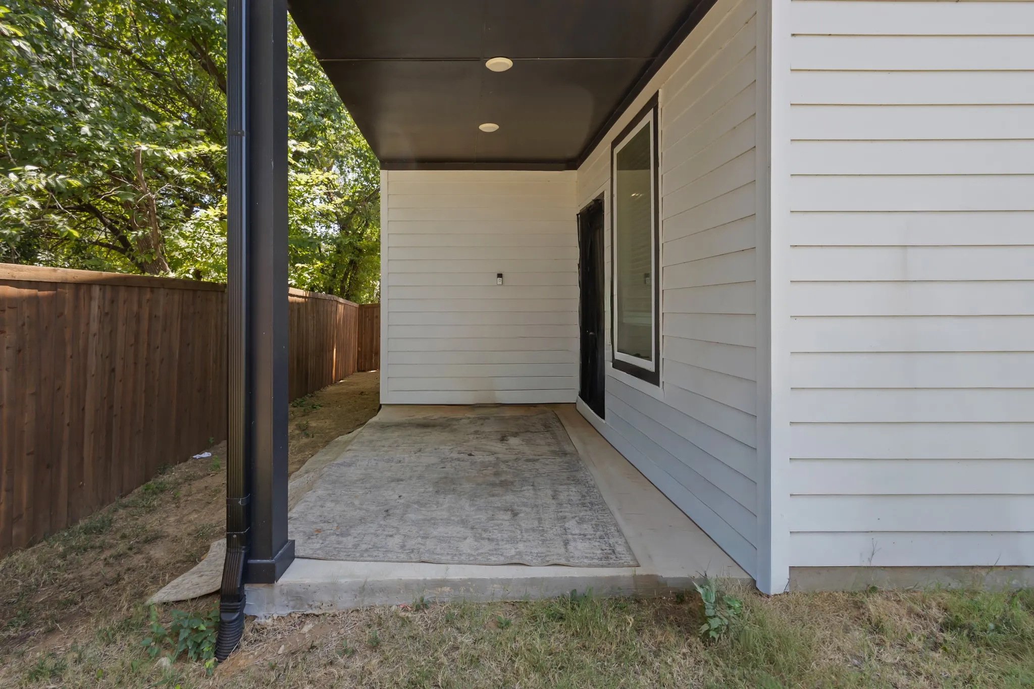 Covered concrete patio with recessed lighting, white horizontal siding, a black exterior door, and a privacy fence