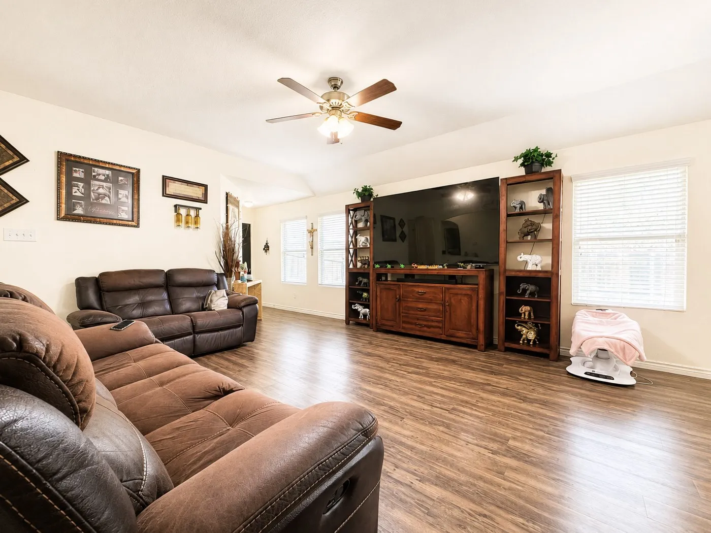 Spacious living area featuring wood-finish flooring, a ceiling fan with integrated lighting, and multiple windows with blinds