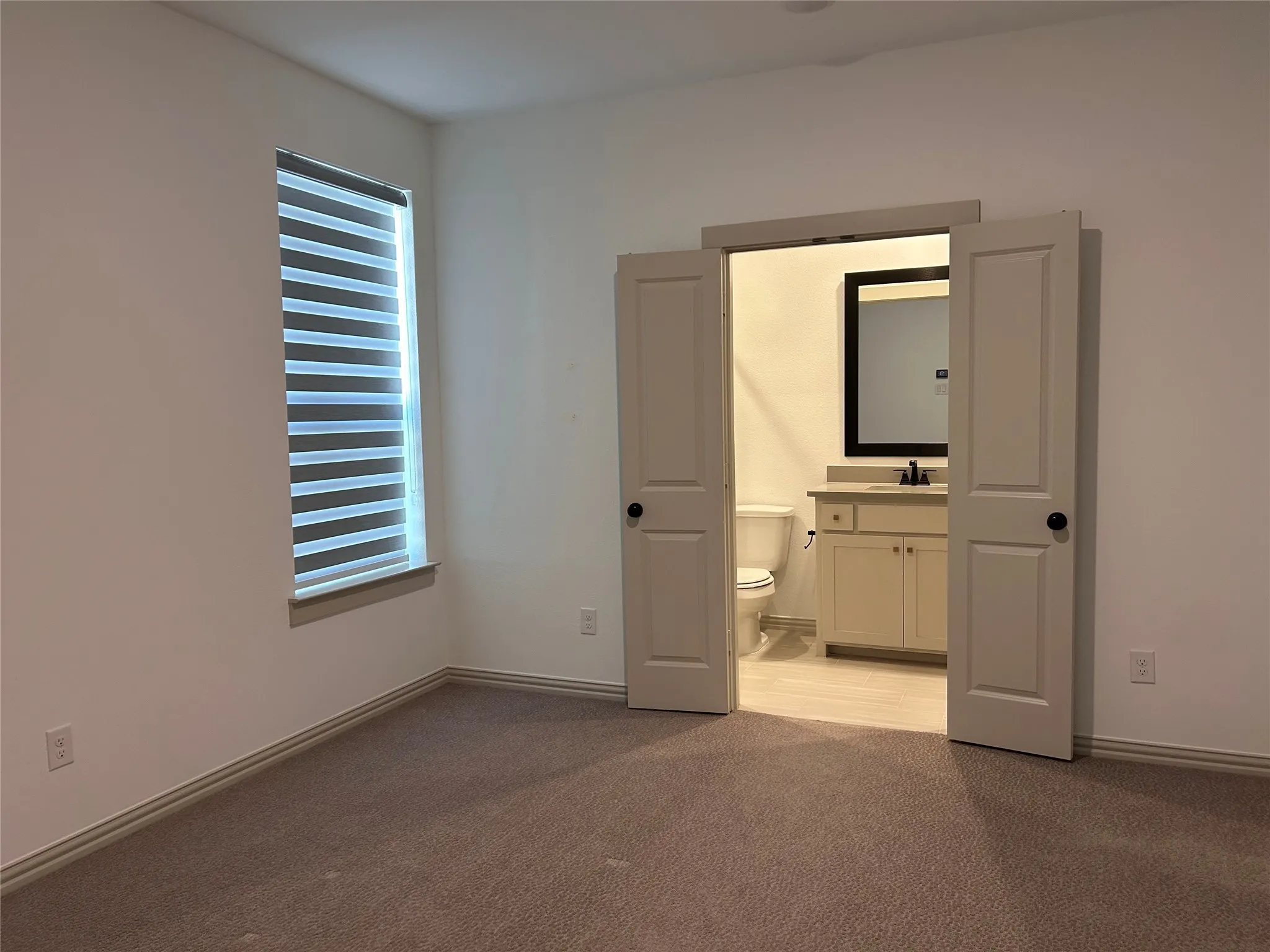 Window with horizontal blinds, carpeted flooring, and double doors opening to a bathroom with a vanity and mirror