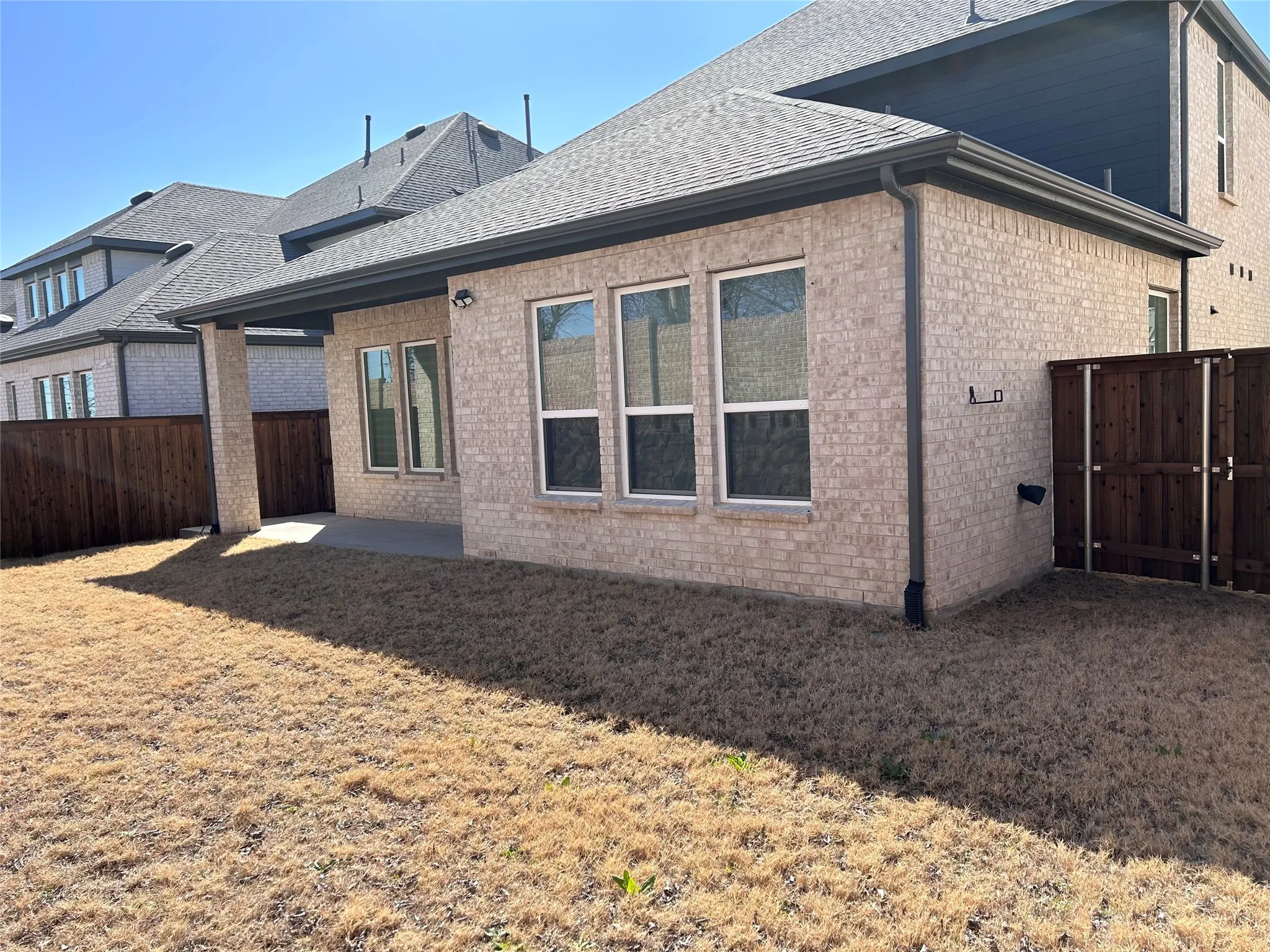 Brick exterior with covered patio, multiple windows, dark wood-tone fencing, and a shingled roof