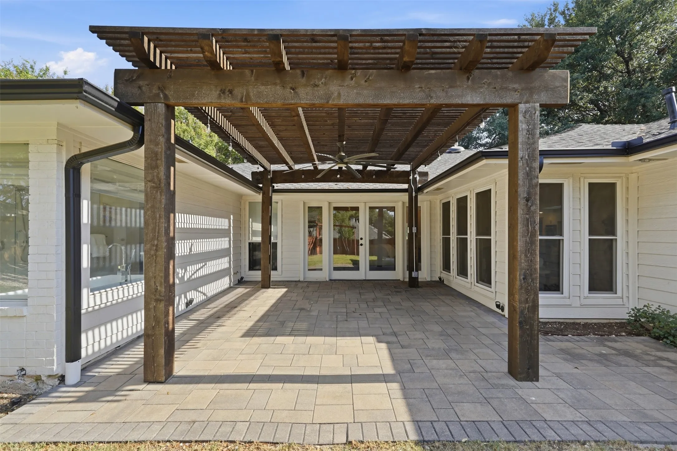 Covered stone paver patio under a dark wood timber pergola with ceiling fan and double glass doors