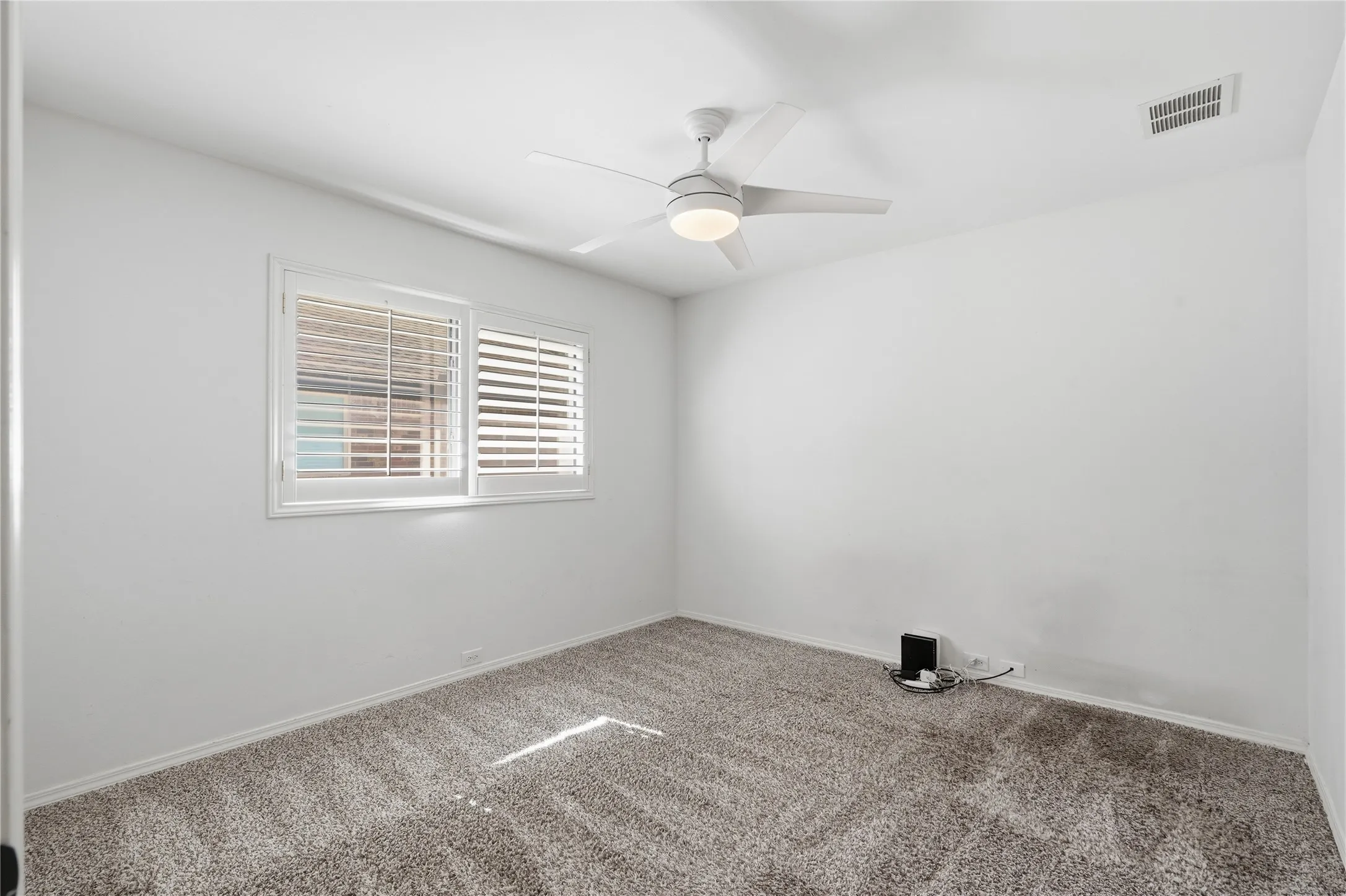 Neutral guest bedroom with plush grey carpet, white ceiling fan, and window plantation shutters