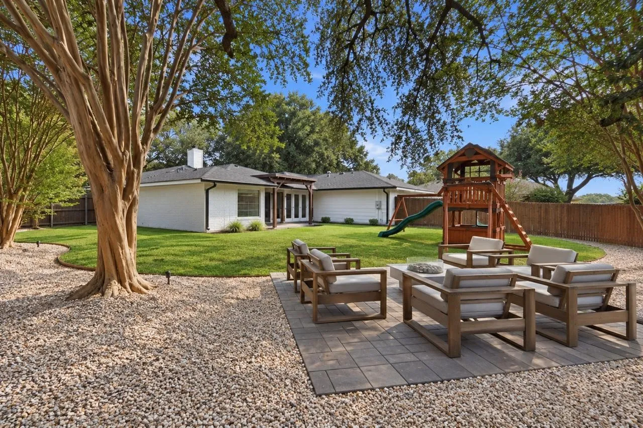 Outdoor stone patio fire pit lounge with six wooden armchairs surrounded by pea gravel and oak trees
