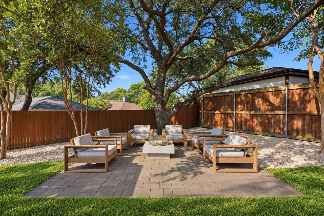 Paver patio with a central fire pit, surrounded by mature trees and a wooden privacy fence, complemented by a gravel perimeter and manicured lawn