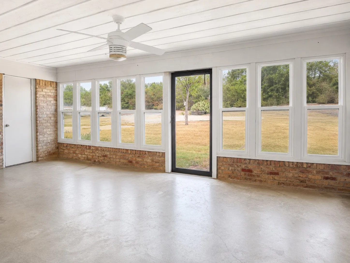 Concrete flooring with brick perimeter walls, multiple windows, a white ceiling fan, and a white paneled ceiling