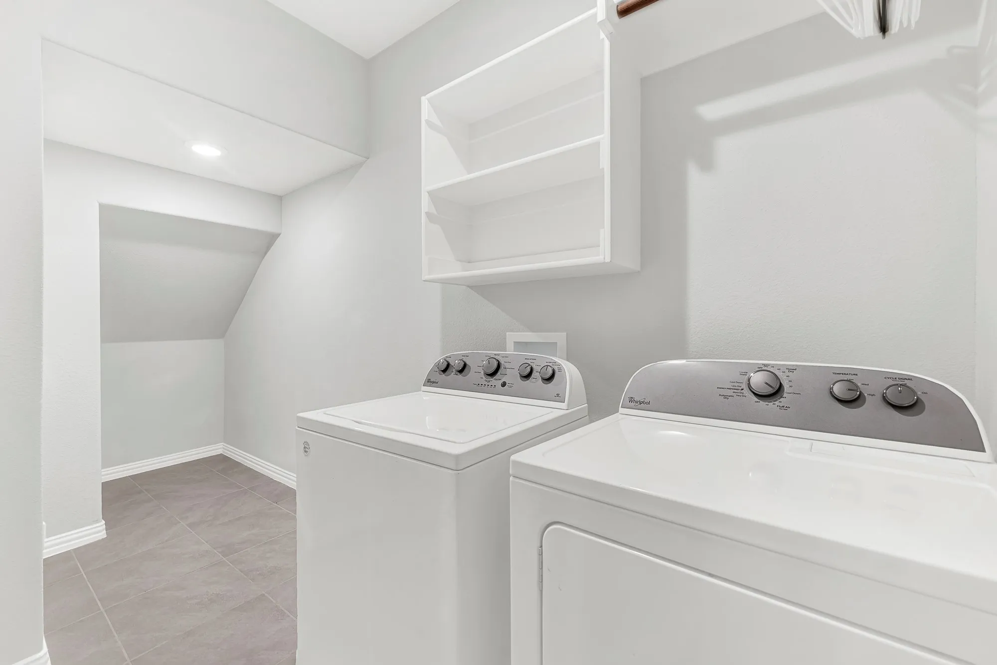 Dedicated laundry room featuring side-by-side washer and dryer units with intuitive control panels, white cabinetry with open shelving for storage, wood-finish flooring, recessed lighting, and direct access to additional storage space