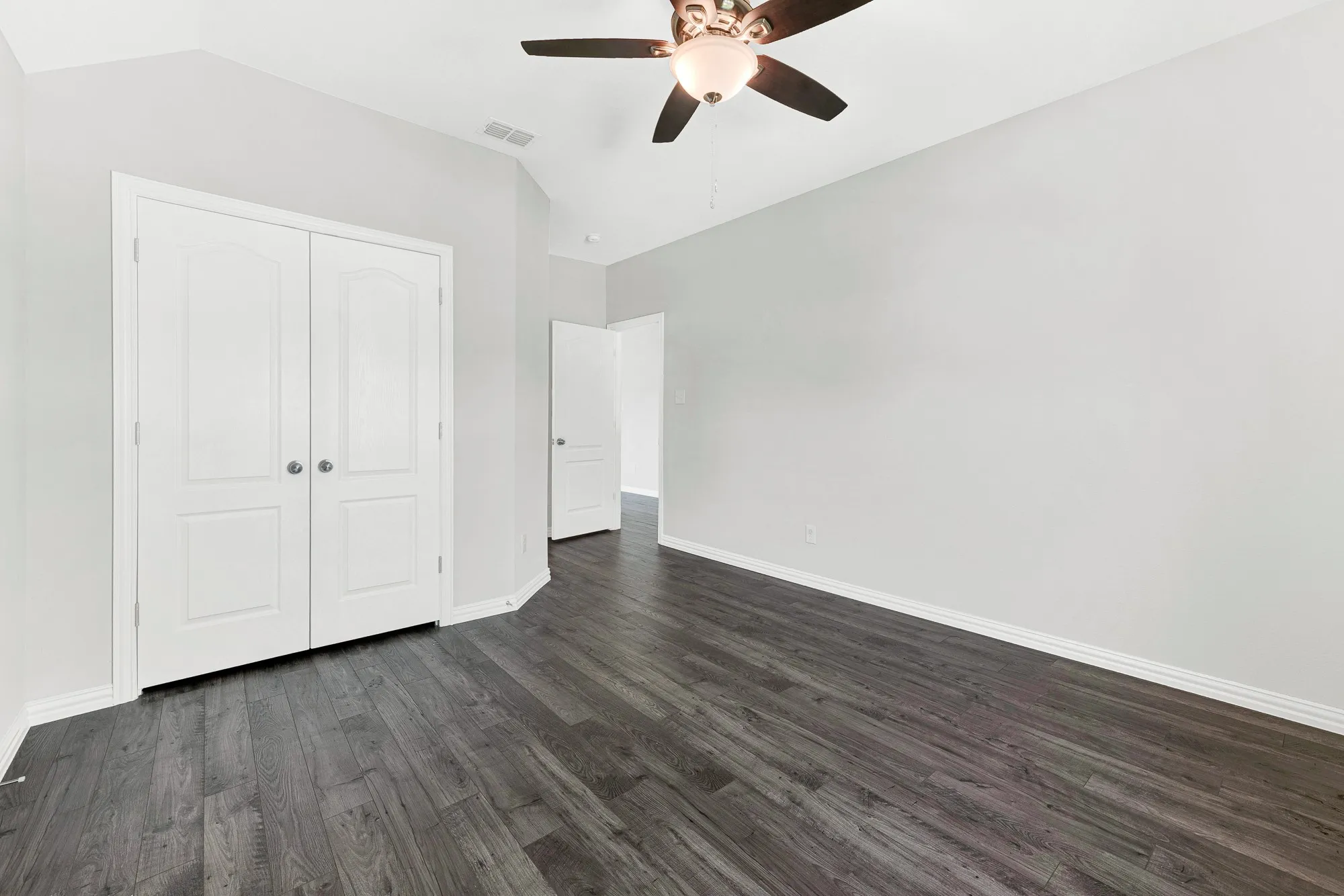 Spacious bedroom featuring dark wood-finish flooring, white panel double-door closet, ceiling fan with integrated lighting, and clean white walls with generous natural light from multiple windows