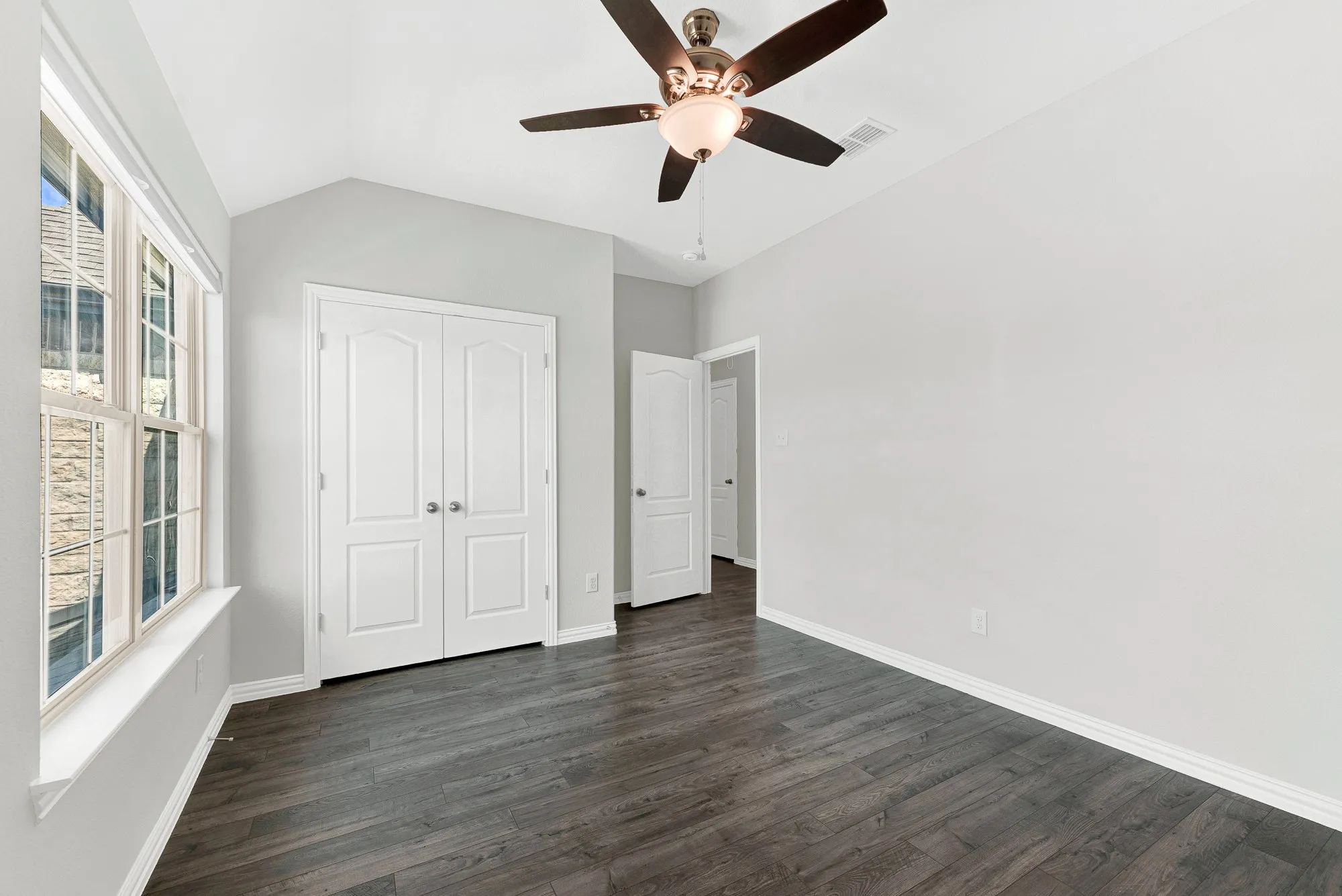 Bright, light-filled bedroom showcasing dark wood-finish flooring, white six-panel closet doors, ceiling fan with integrated lighting, and expansive windows providing natural illumination throughout the space