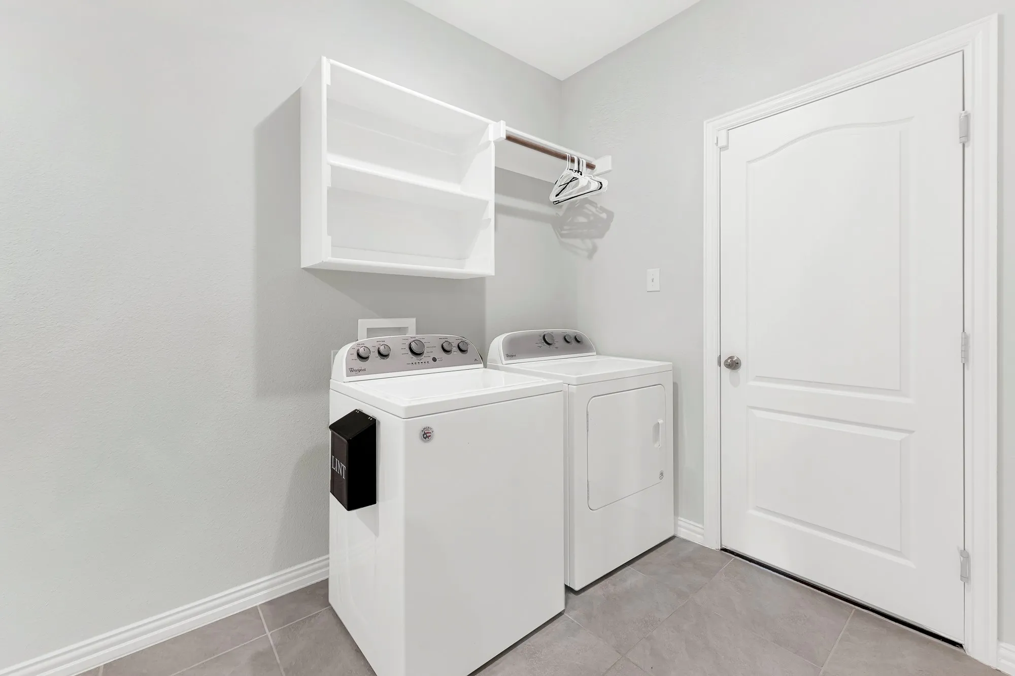 Dedicated laundry room featuring side-by-side washer and dryer in white, upper cabinetry with glass-front doors and shelving, brushed nickel hardware, and neutral tile flooring