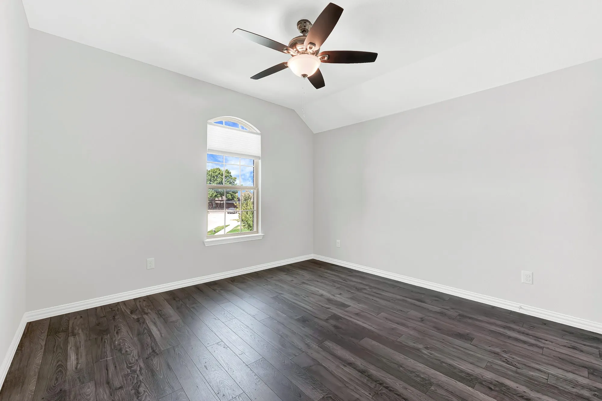 Bright corner bedroom showcasing dark wood-finish flooring, clean white walls, and abundant natural light from an arched window