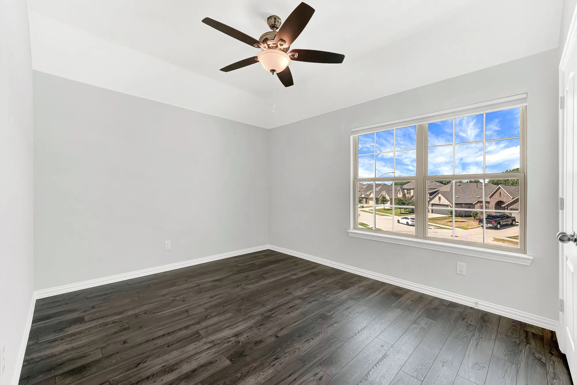 Bright corner bedroom showcasing dark wood-finish flooring, ceiling fan with integrated lighting, and expansive multi-pane windows framing natural light and neighborhood scenery