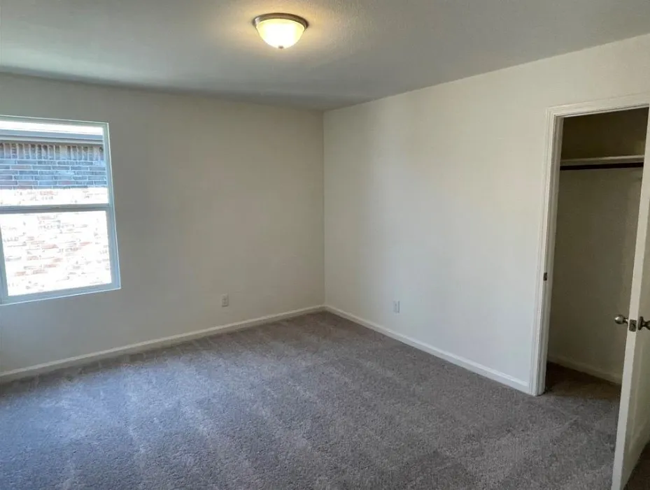 Wall-to-wall carpeting in a neutral tone, ceiling-mounted light fixture, white interior walls, and a window with a brick exterior visible