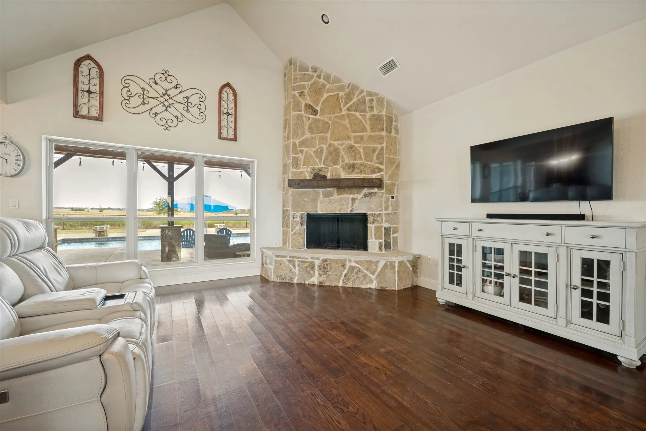 Vaulted ceiling, stone-clad wood burning fireplace with wood mantle, dark wood-finish flooring, and large windows overlooking an outdoor pool