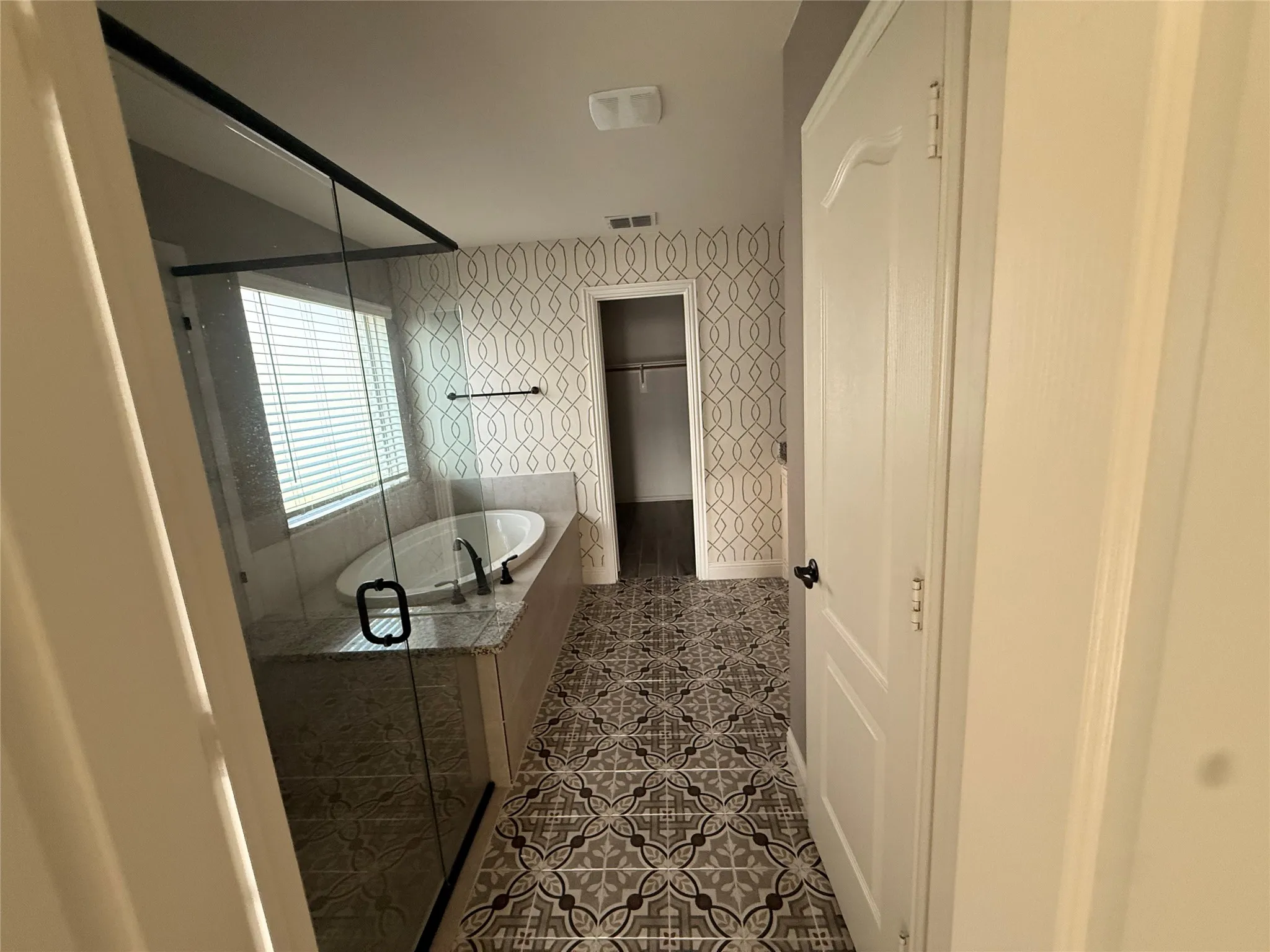 Patterned tile flooring, recessed bathtub, full-height window with blinds, glass-enclosed shower, geometric patterned wall