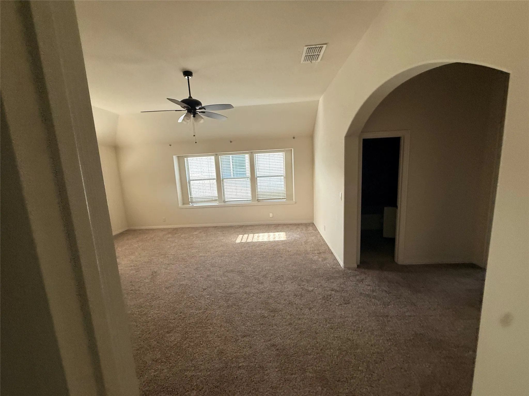 Spacious room featuring carpeted flooring, multiple windows with blinds, a ceiling fan, and an arched doorway