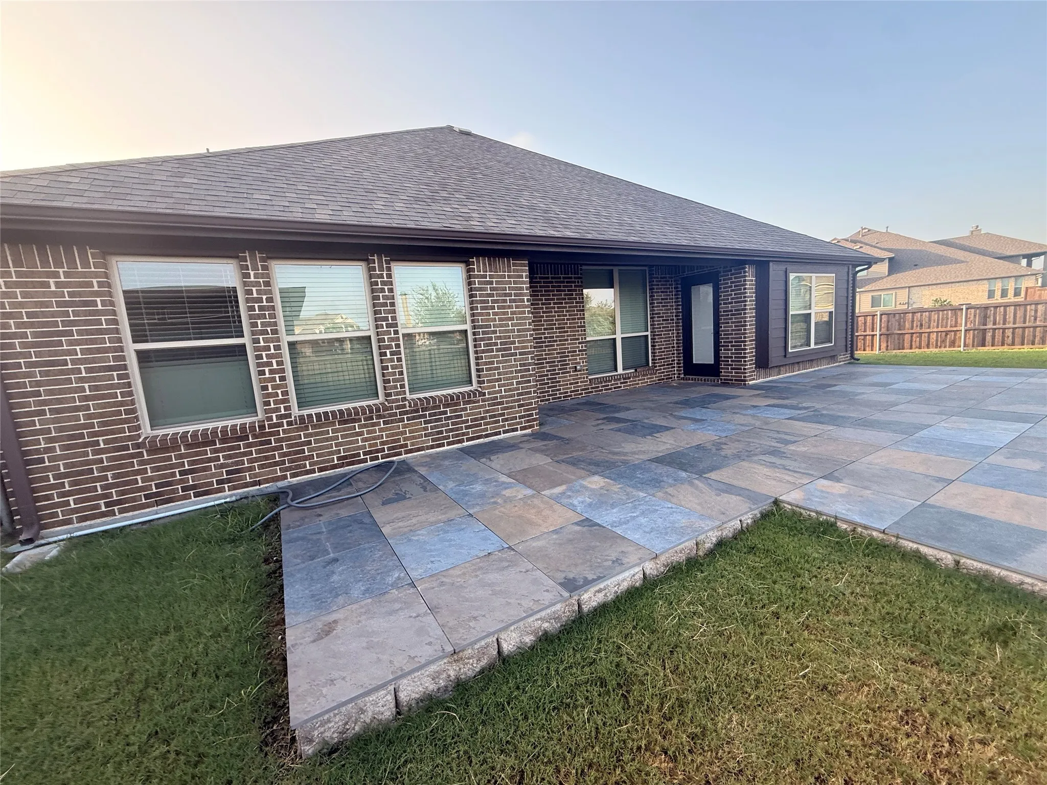 Expansive backyard patio featuring large format pavers, brick and siding exterior, multiple windows, and a dark shingled roof