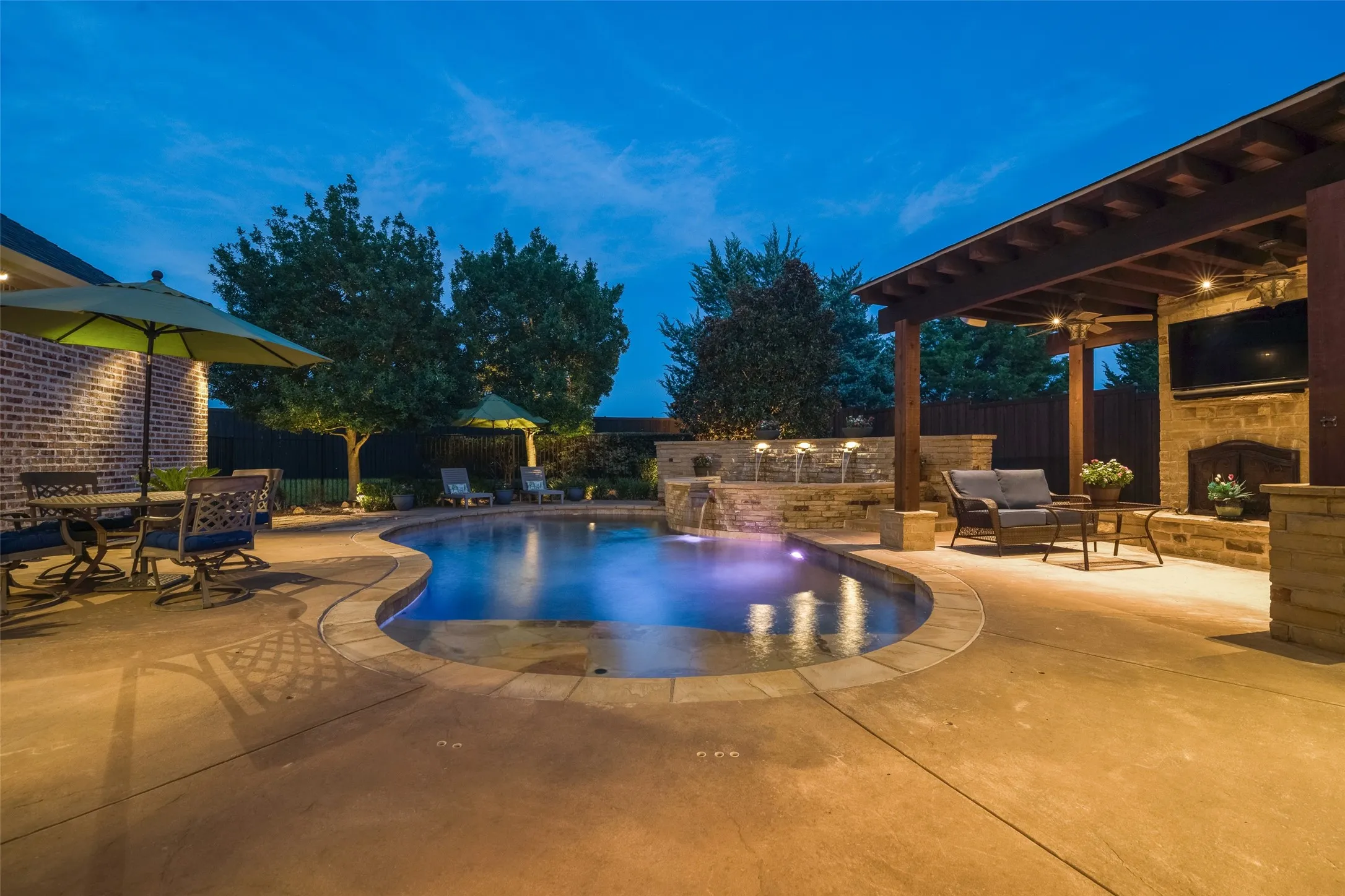 Curved swimming pool with integrated water features, stone-clad outdoor fireplace, wood pergola, and extensive concrete patio