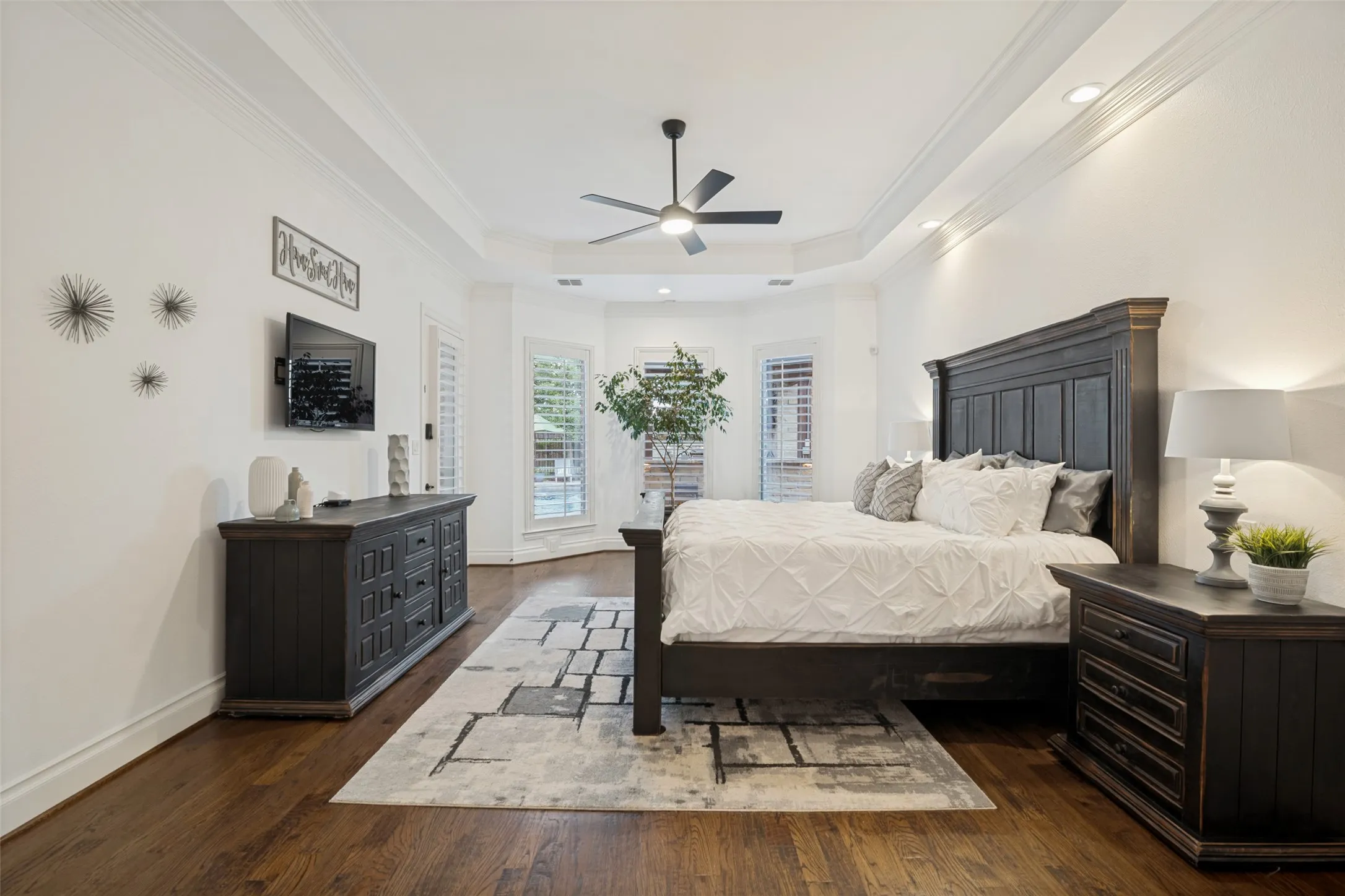 Hardwood flooring, tray ceiling with recessed lighting, ceiling fan, bay window with plantation shutters, and crown molding