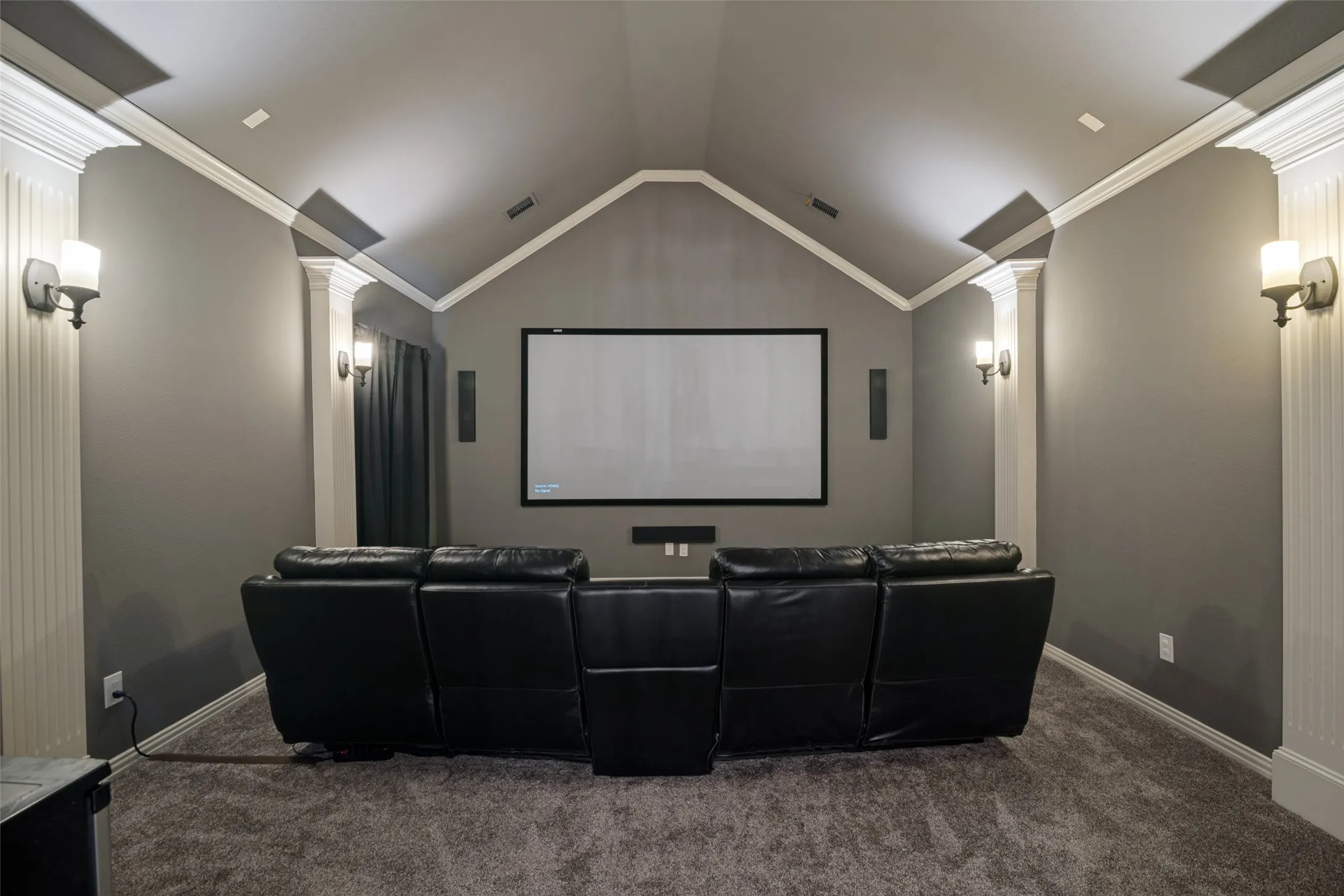 Vaulted ceiling with crown molding, wall-mounted sconces, built-in projection screen, integrated speakers, and carpeted flooring