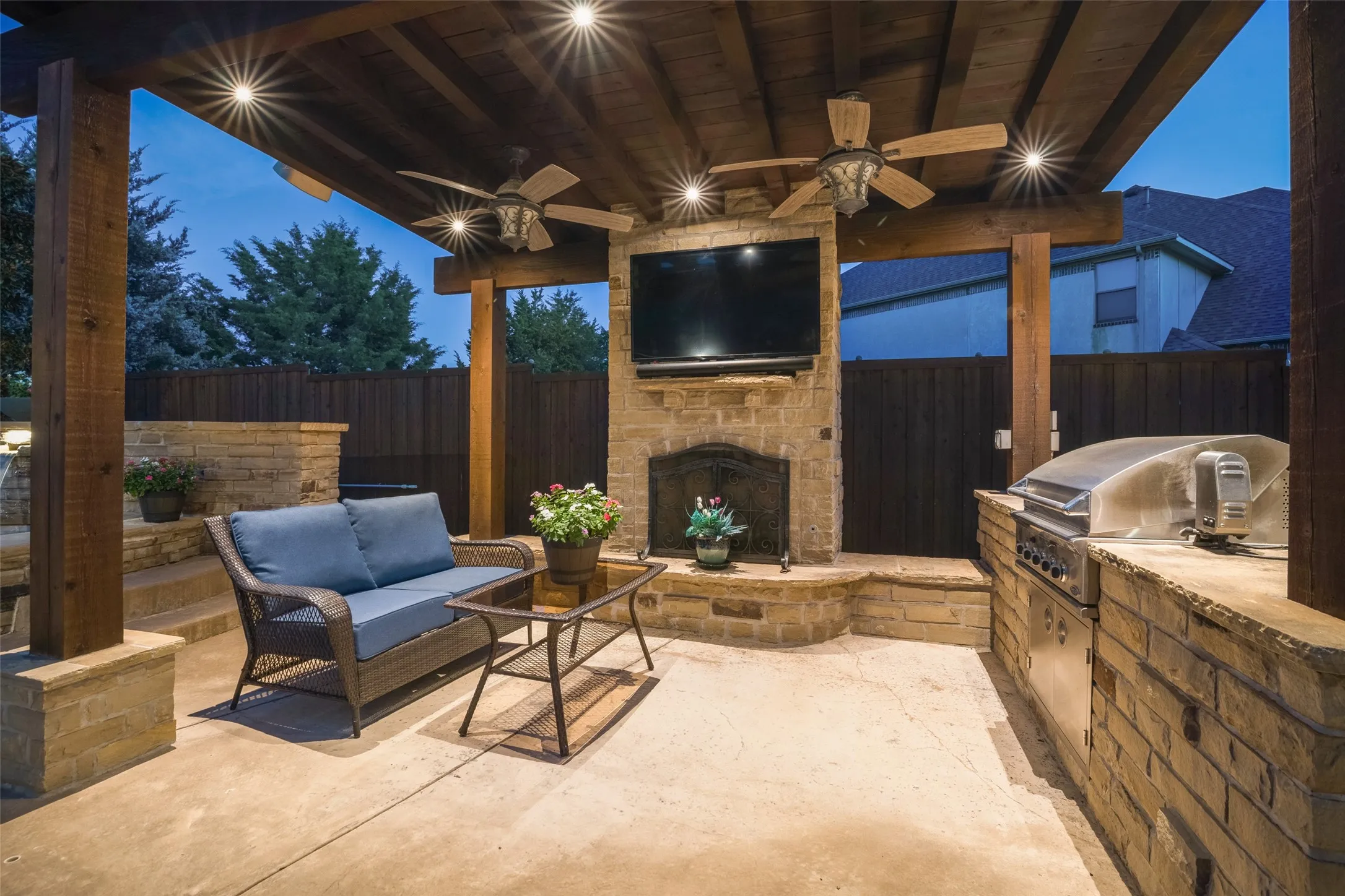 Covered patio with integrated lighting, exposed wood-beam ceiling, stone-clad fireplace, built-in outdoor grill, and two ceiling fans